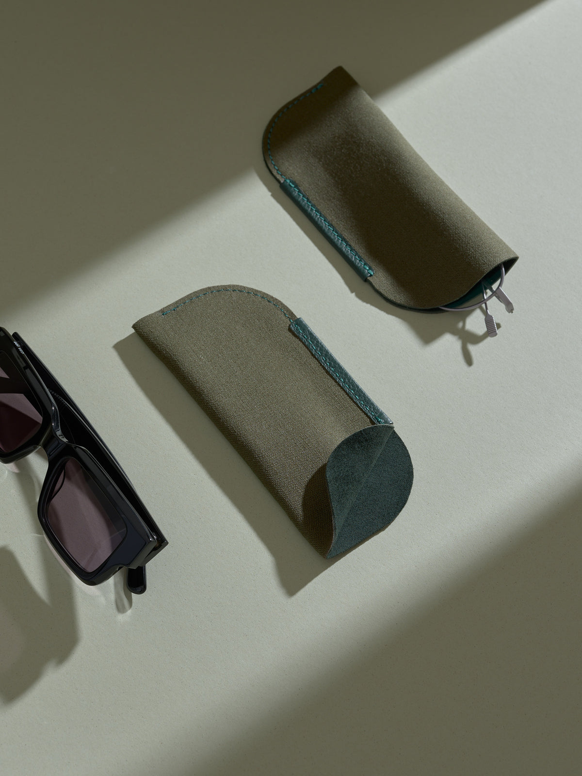 Two separate soft olive-green fabric sleeves, designed to hold glasses, are displayed on a light green surface, next to a pair of black rectangular sunglasses.