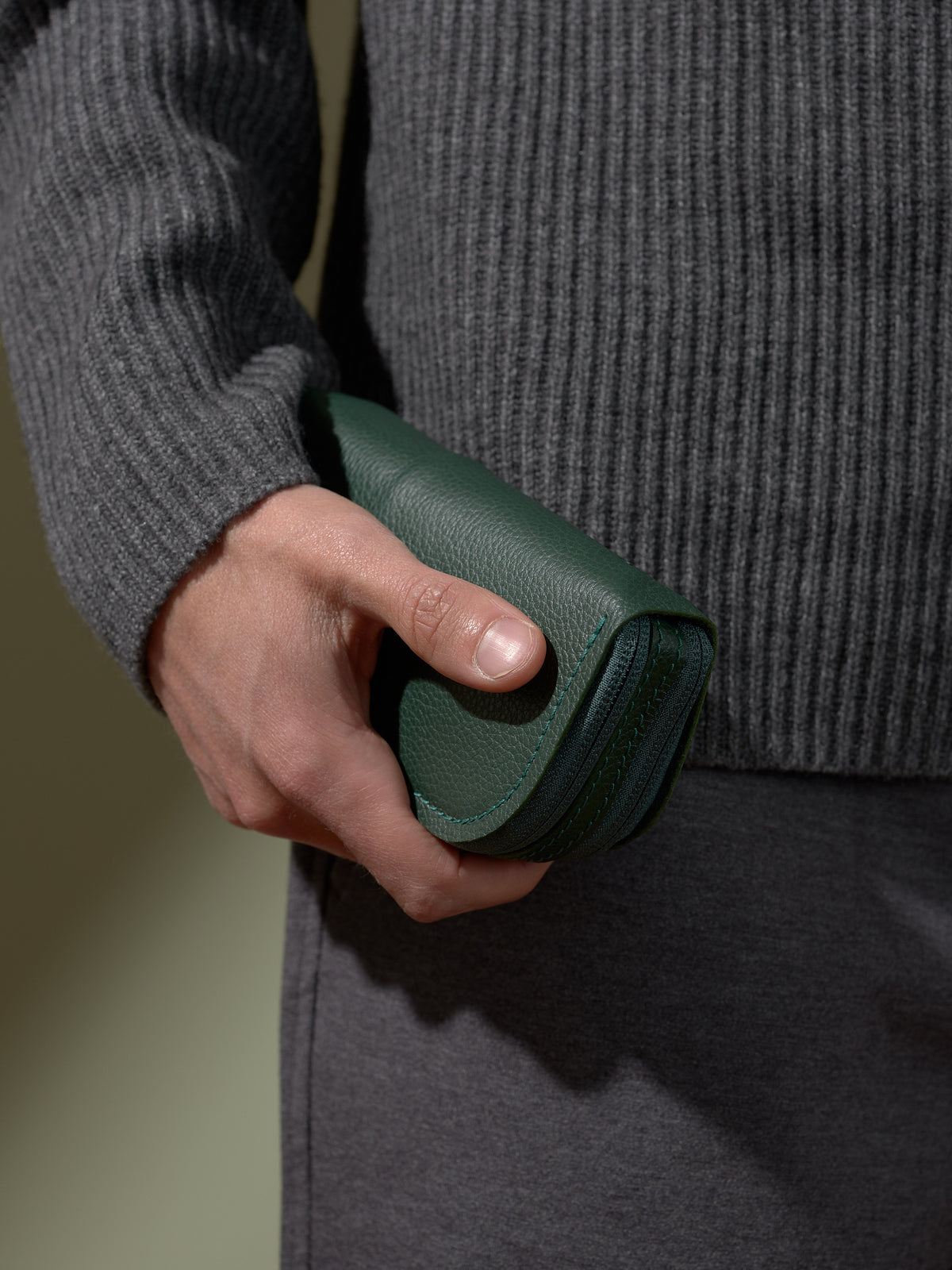 A person's hand holding the closed, pebbled green Capra leather double glasses case vertically, set against a background of a dark gray sweater and trousers.