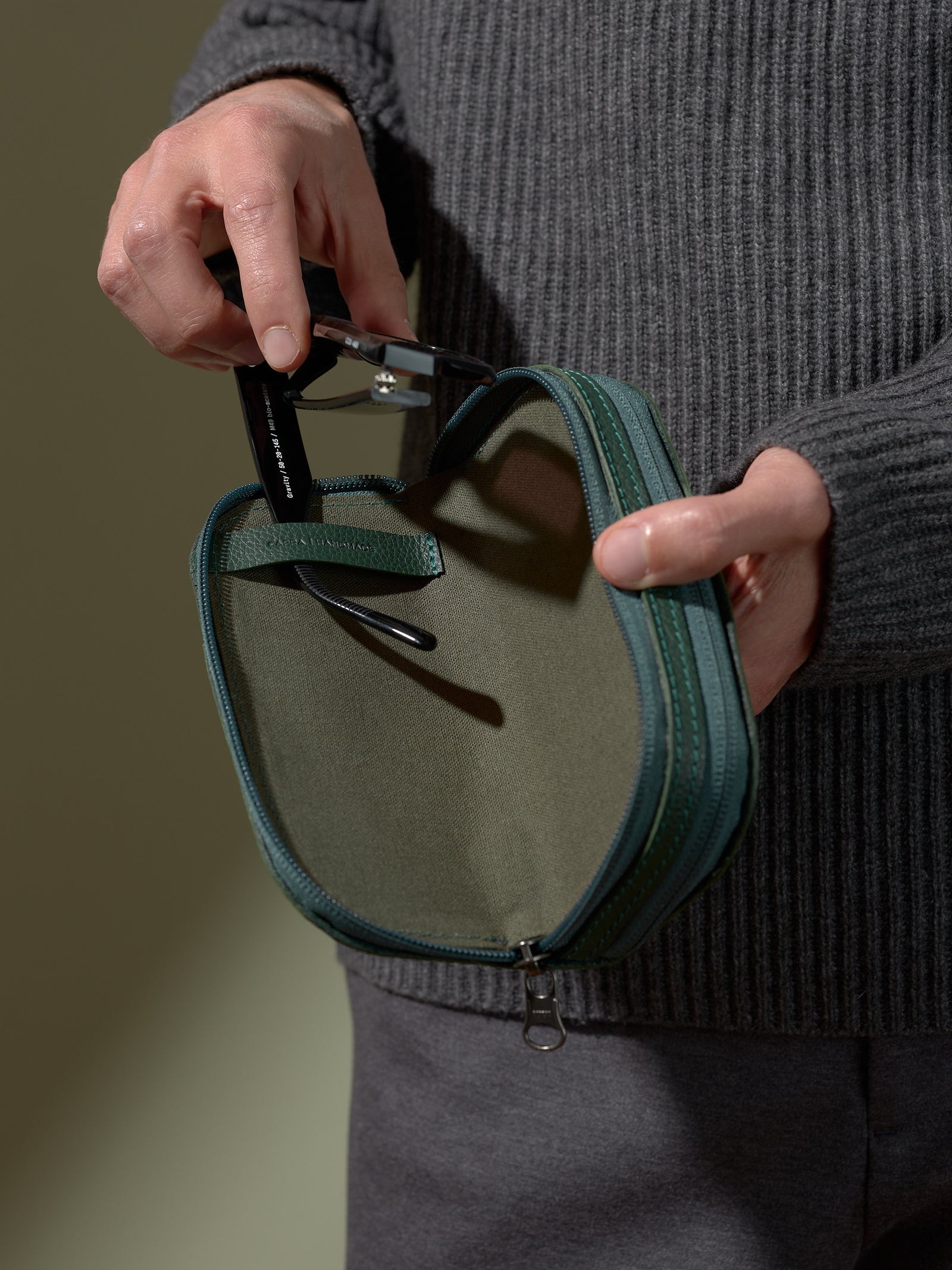 Close-up of a person's hands inserting a pair of black-framed glasses into the open, zipped green leather case, while wearing a dark gray sweater.