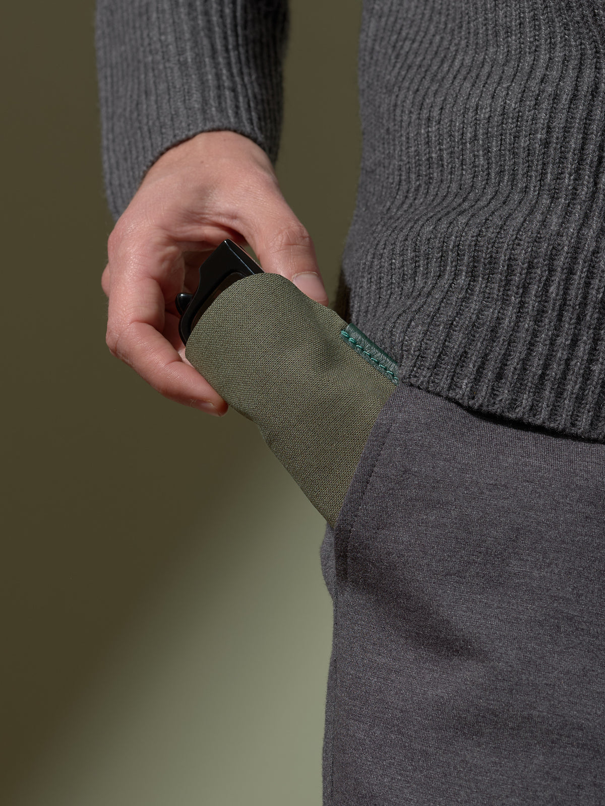 Close-up of a person's hand inserting the soft olive-green fabric inner sleeve of the glasses case, with a pair of black sunglasses inside, into the pocket of dark gray trousers.