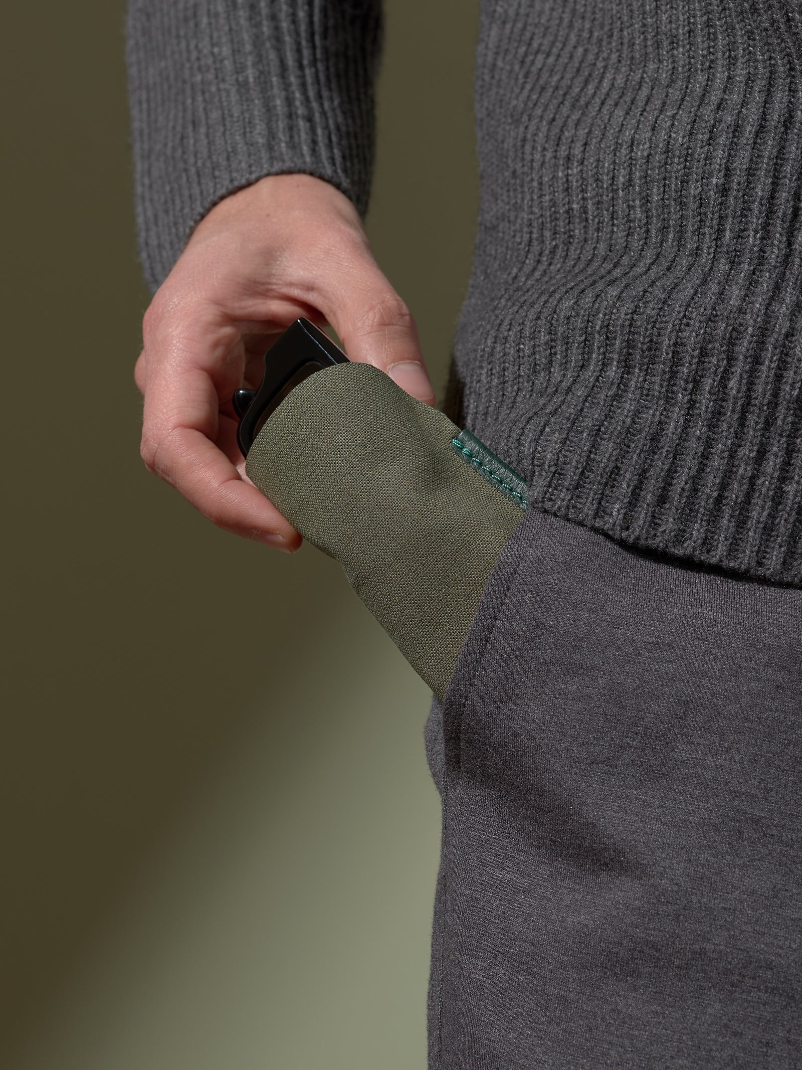 Close-up of a person's hand inserting the soft olive-green fabric inner sleeve of the glasses case, with a pair of black sunglasses inside, into the pocket of dark gray trousers.