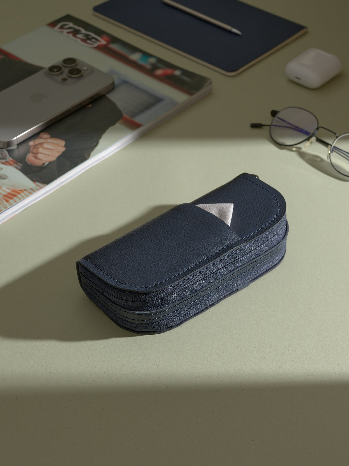 The closed, navy blue Capra leather double glasses case resting on a light green desk, with a small white cleaning cloth sticking out of the exterior pocket. A phone, notepad, and glasses are visible in the background.