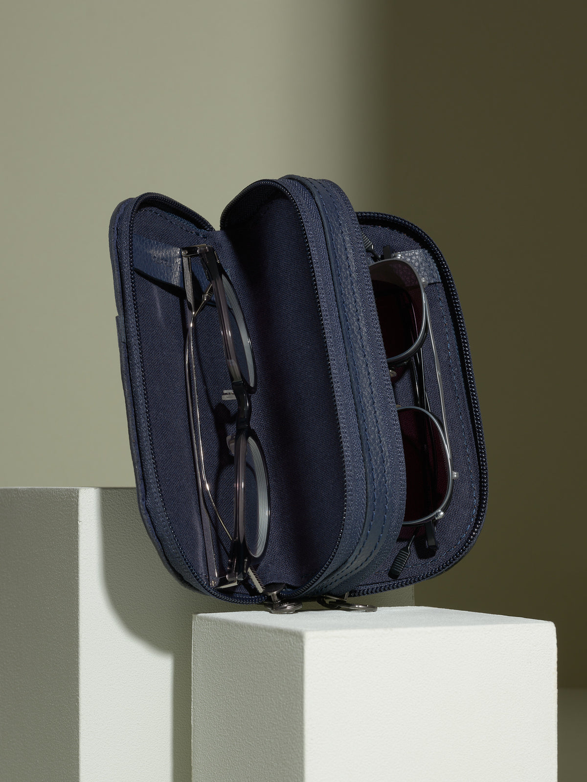 The opened, navy blue leather double glasses case standing upright on white platforms, displaying two pairs of glasses stored inside: clear glasses on the left and sunglasses on the right.