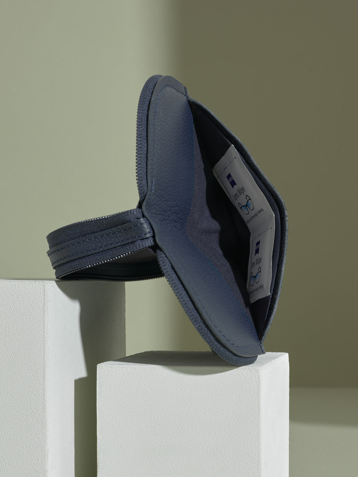 The opened, navy blue leather glasses case standing upright on white platforms, showing the dark blue leather interior and a white object (possibly an AirTag or earbud case) in a small interior pocket.