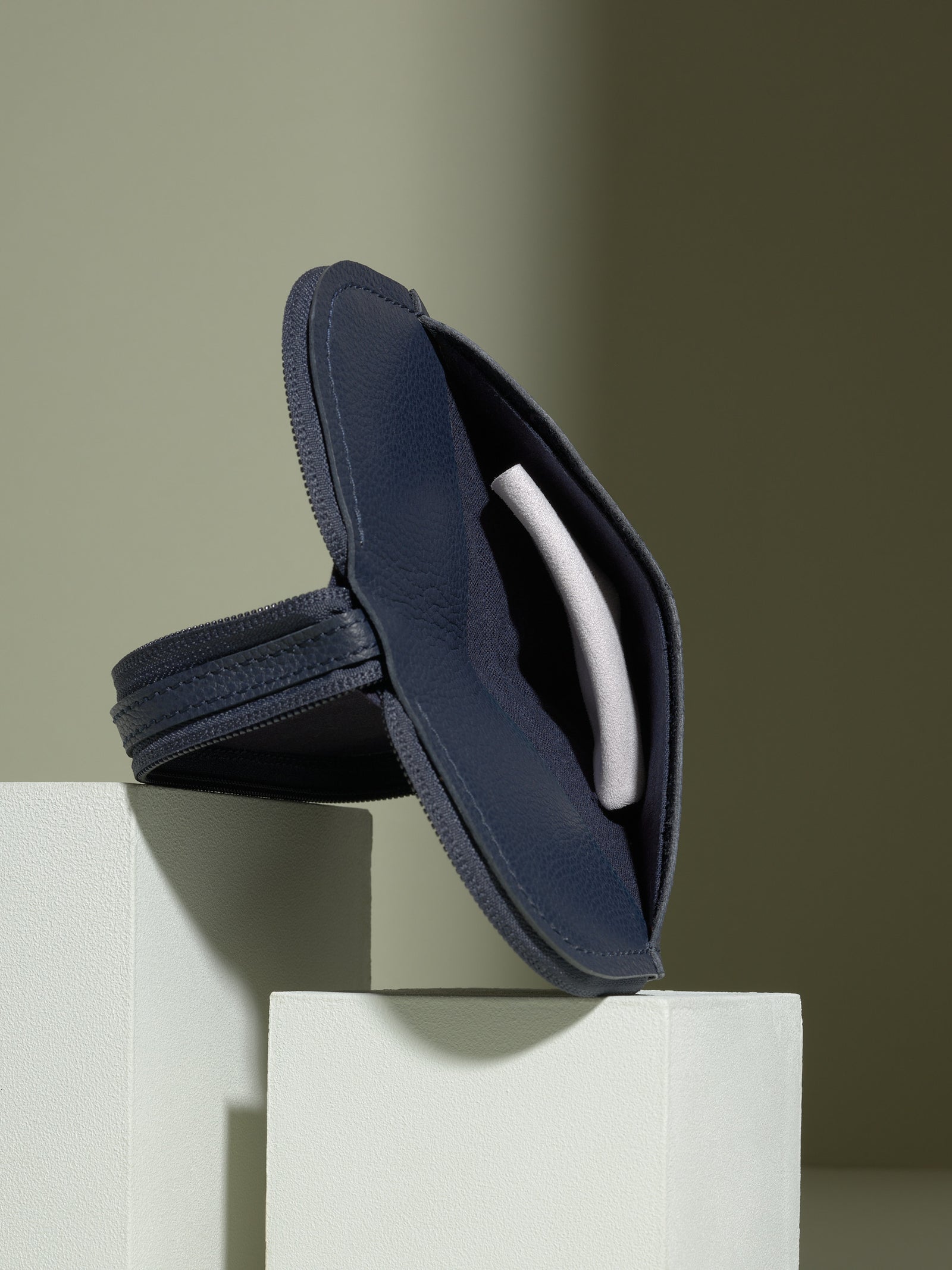 The opened, navy blue leather glasses case standing upright on white platforms, showing the dark blue leather interior and a white object (possibly an AirTag or earbud case) in a small interior pocket.