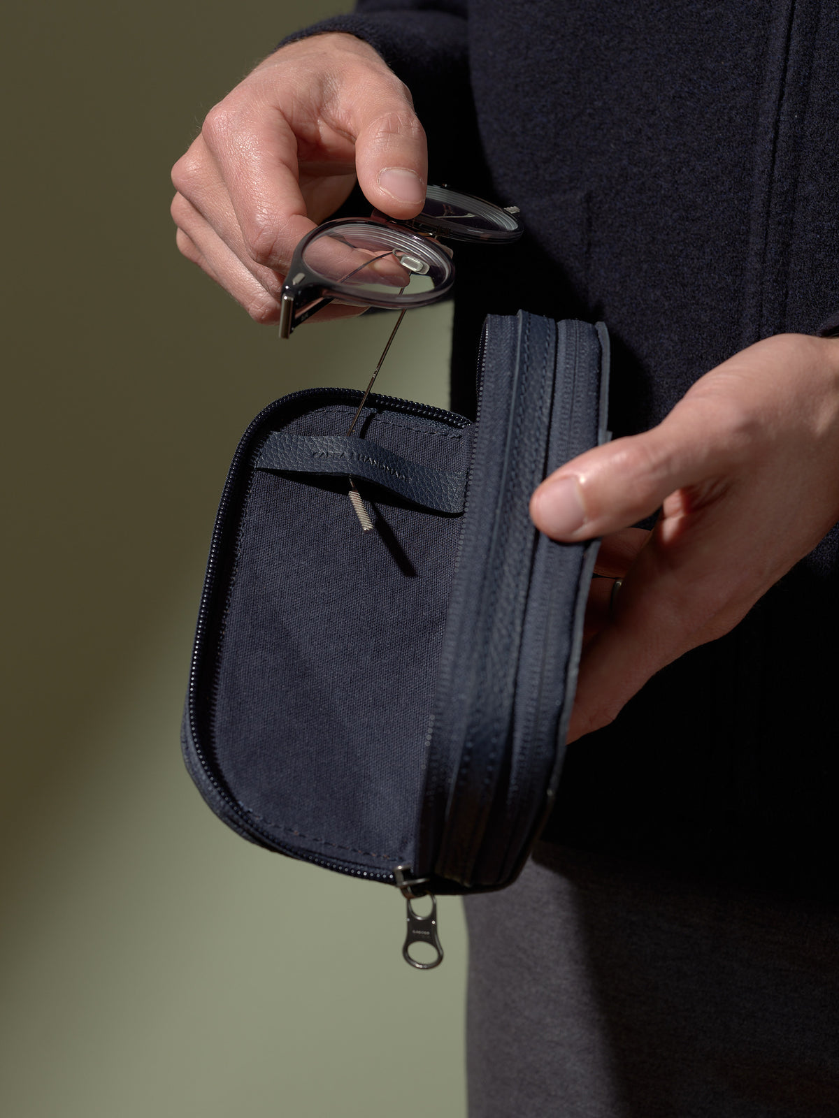 A person's hands holding the opened, navy blue leather glasses case and removing a pair of clear glasses from the soft inner compartment.