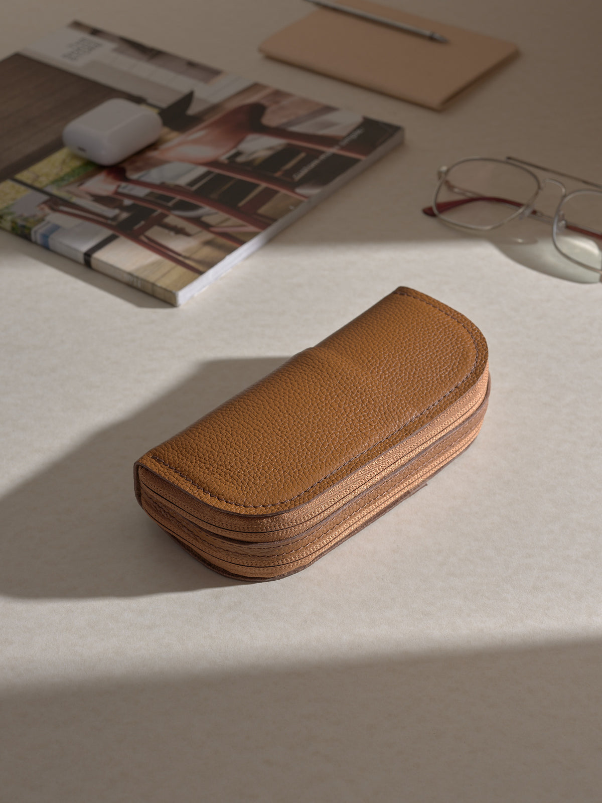 The tan leather double glasses case lying on a light-colored desk surface, surrounded by other items like a magazine, a notepad, a pen, and a pair of glasses.