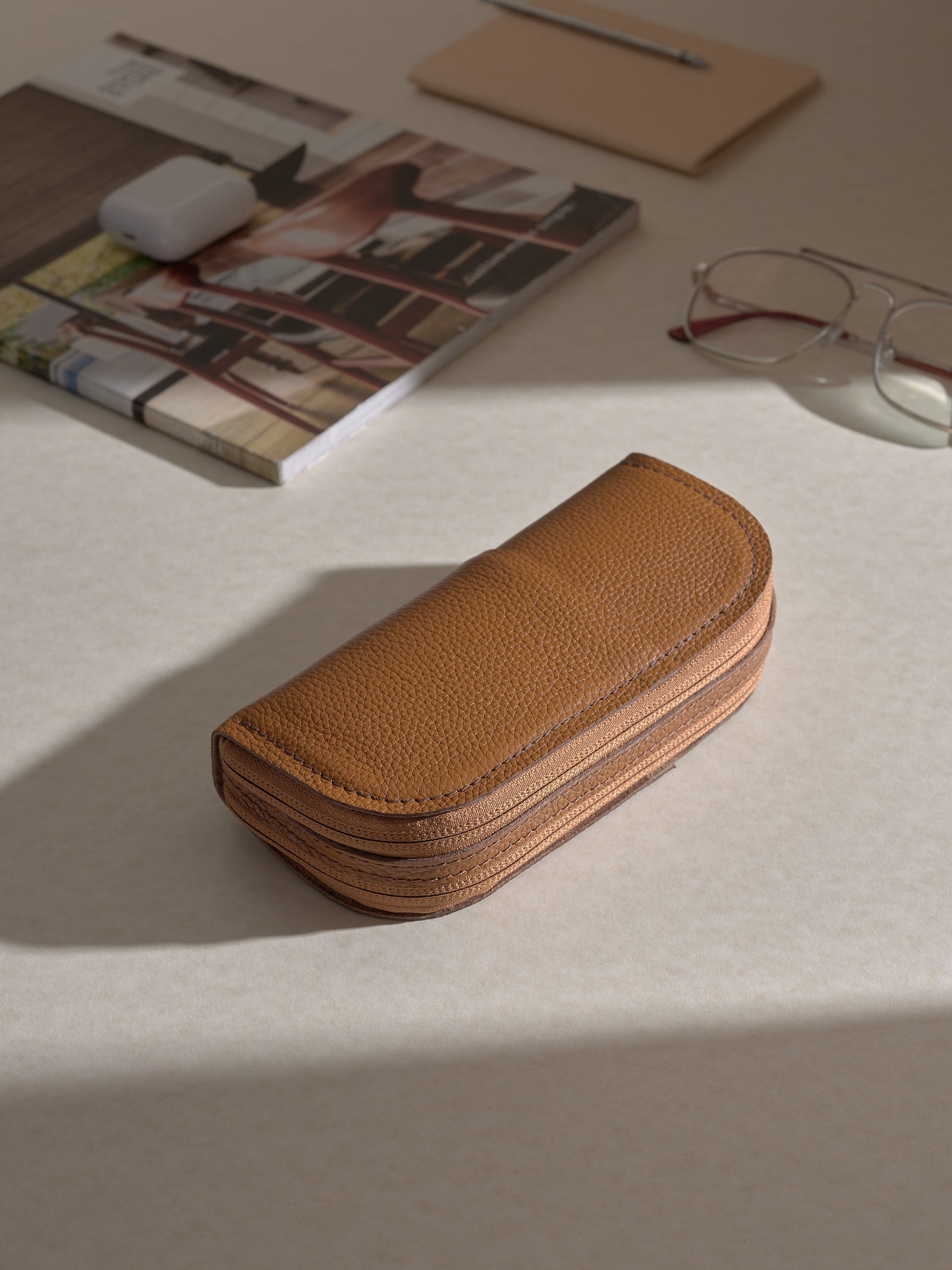 The tan Capra leather double glasses case resting horizontally on two white platforms, with a pair of silver-framed glasses positioned behind it on the upper platform.