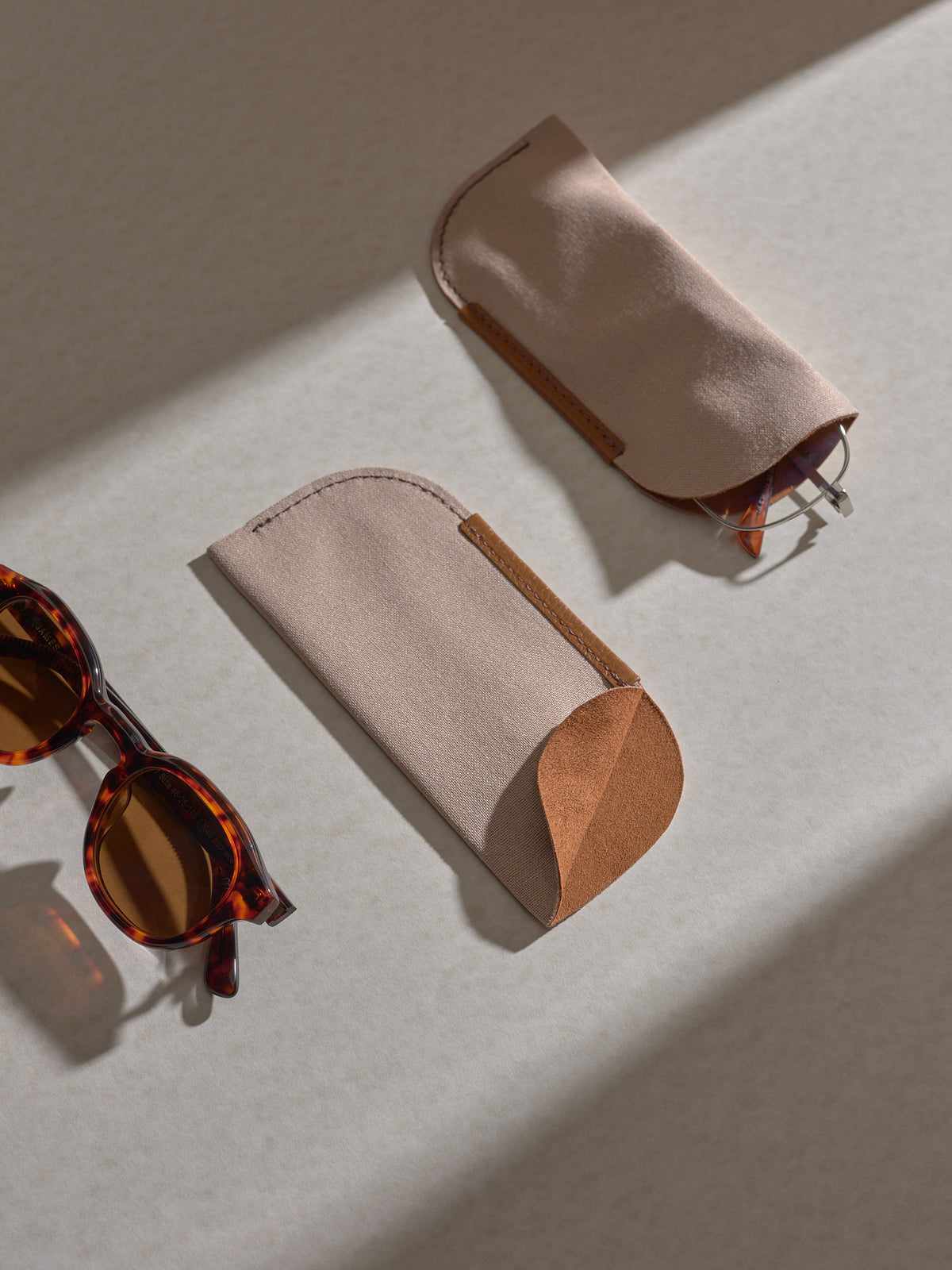 Two separate soft fabric sleeves, which hold the glasses within the main case, are shown on a light surface, with a pair of tortoise-shell sunglasses next to them.