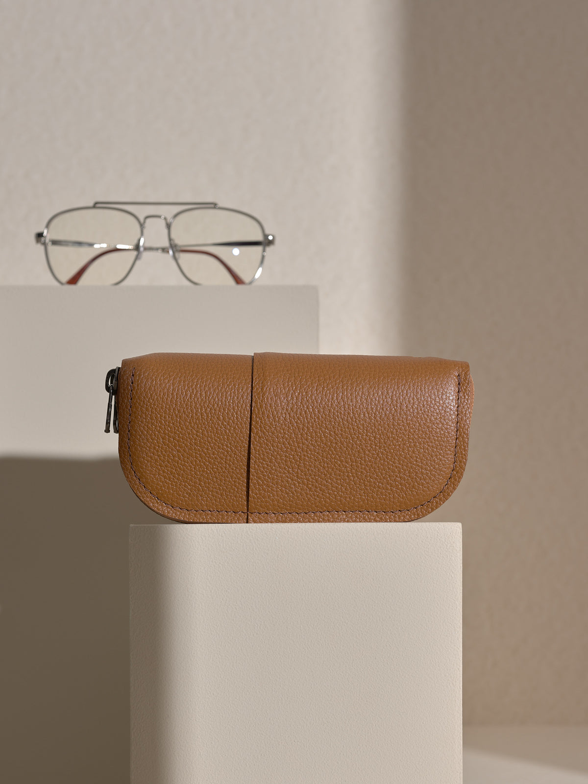 The tan Capra leather double glasses case resting horizontally on two white platforms, with a pair of silver-framed glasses positioned behind it on the upper platform.
