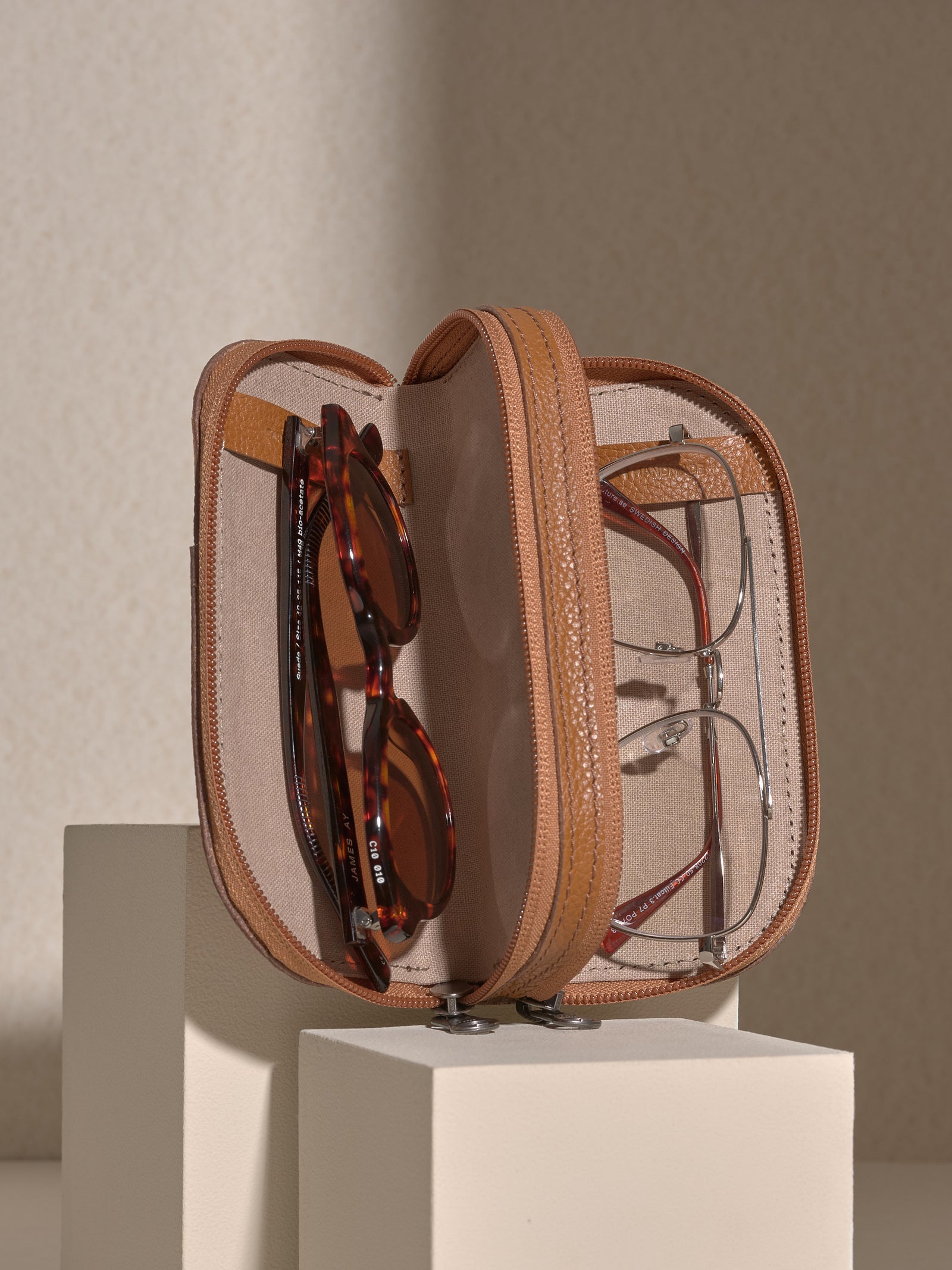 The zipped tan leather double glasses case opened to show two pairs of glasses stored inside: tortoise-shell sunglasses in one compartment and silver-framed clear glasses in the other.