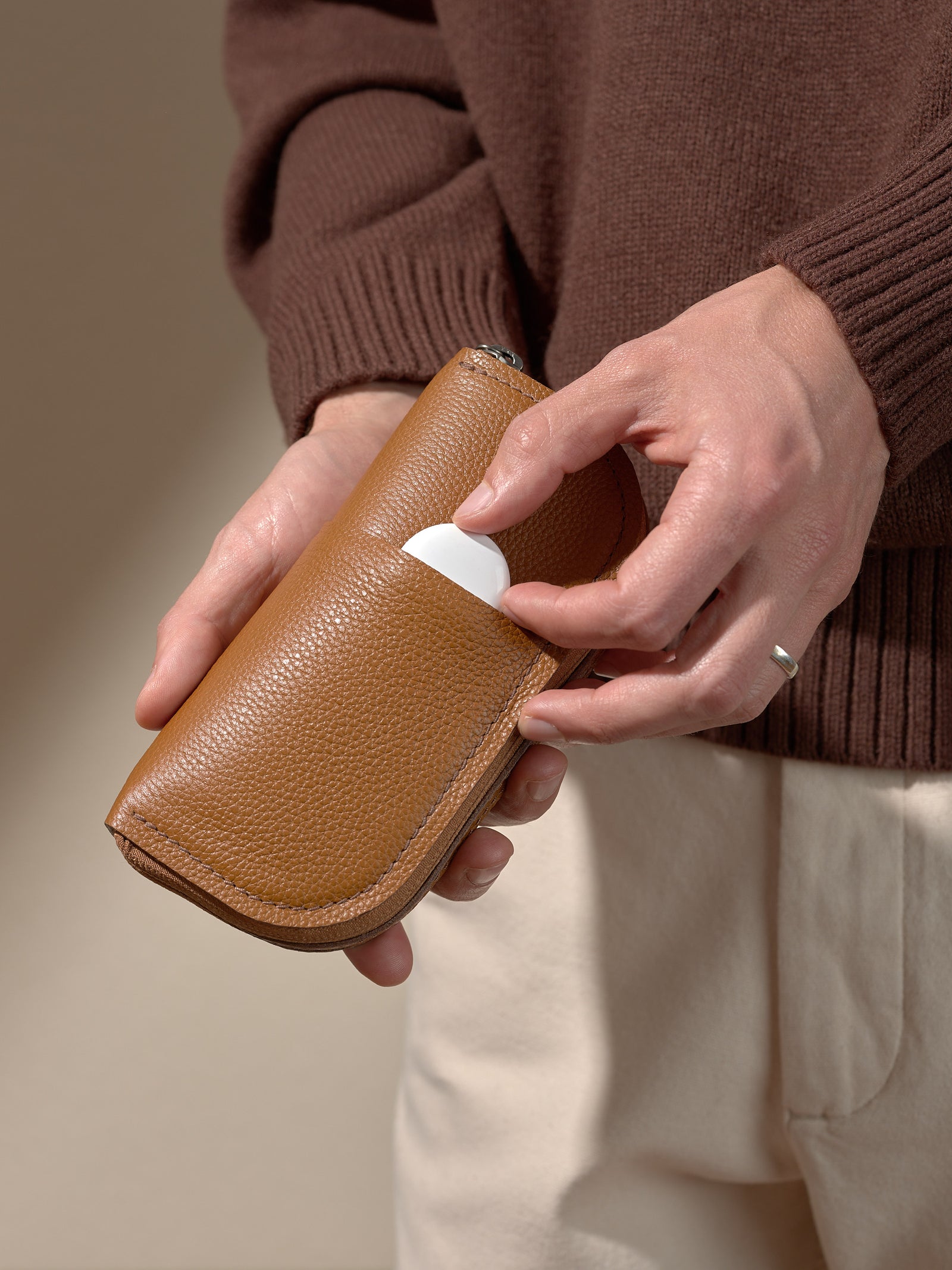 A person's hands holding the closed, textured tan leather glasses case and placing a small, white, circular object (possibly an AirTag or earbud case) into a small exterior slip pocket