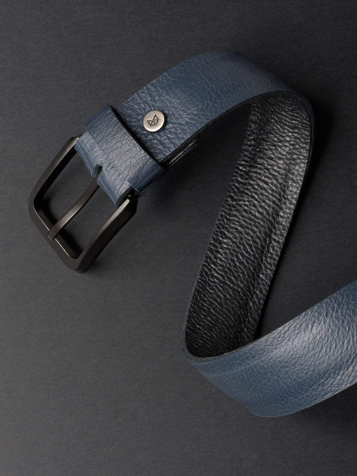 Close-up of a blue grain leather belt with a matte black buckle and a silver Capra Leather logo stud.