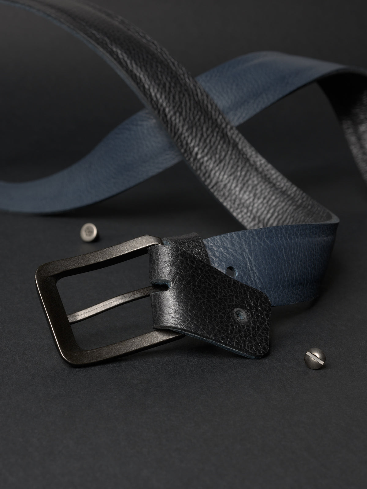 Disassembled reversible leather belt showing the screw mechanism, black and navy leather, and black buckle.