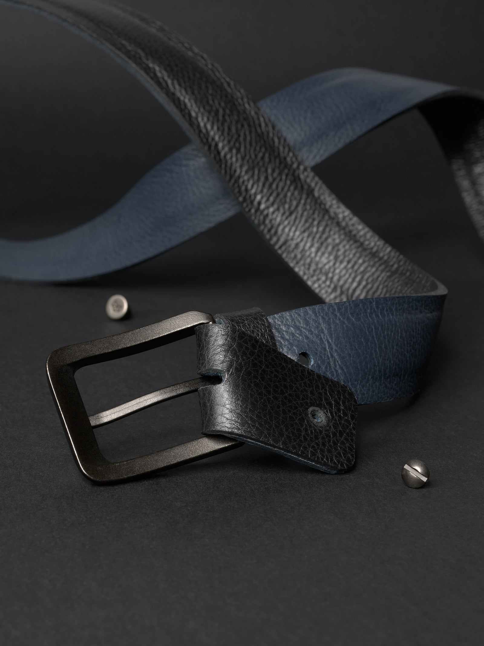 Disassembled reversible leather belt showing the screw mechanism, black and navy leather, and black buckle.