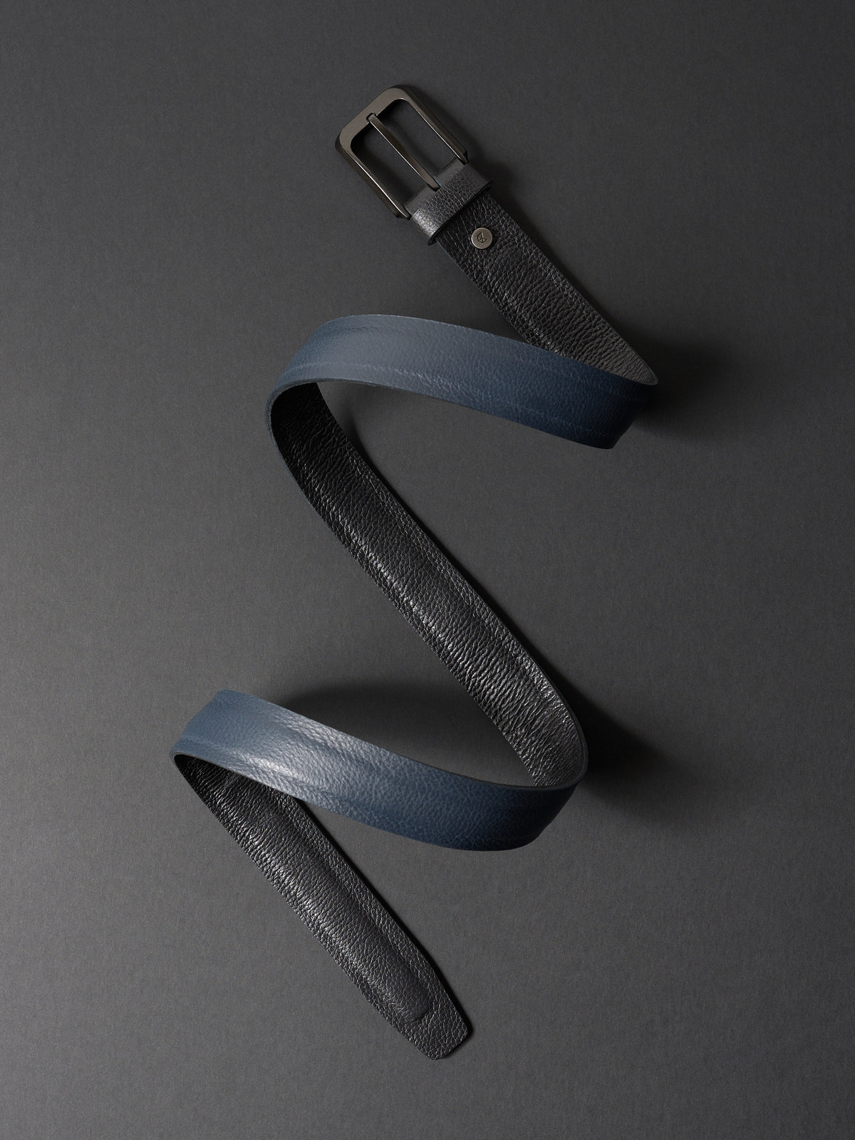 Overhead view of a coiled double-sided 32mm leather belt in black and navy on a dark gray background.