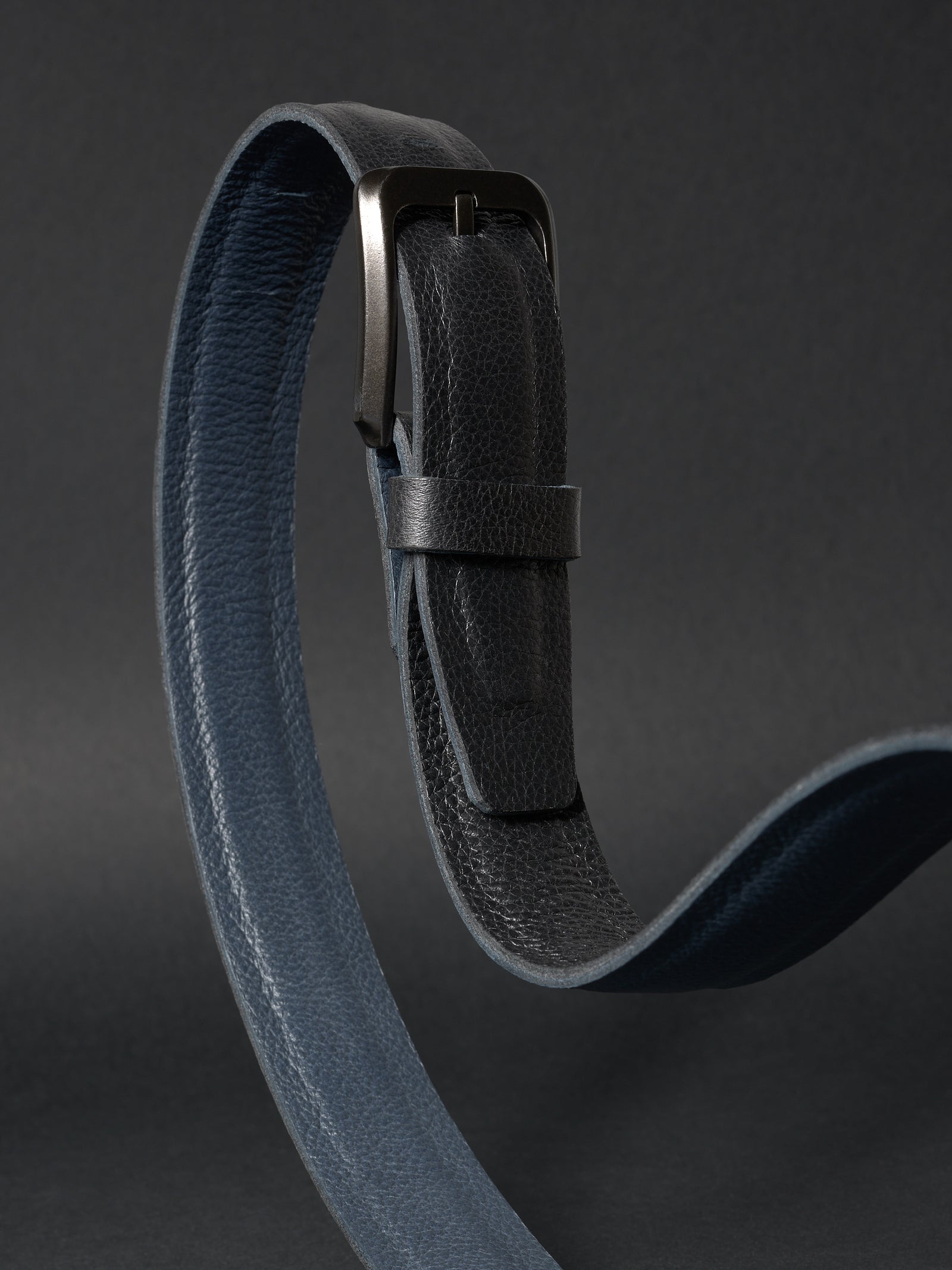 Overhead view of a coiled double-sided 32mm leather belt in black and navy on a dark gray background.