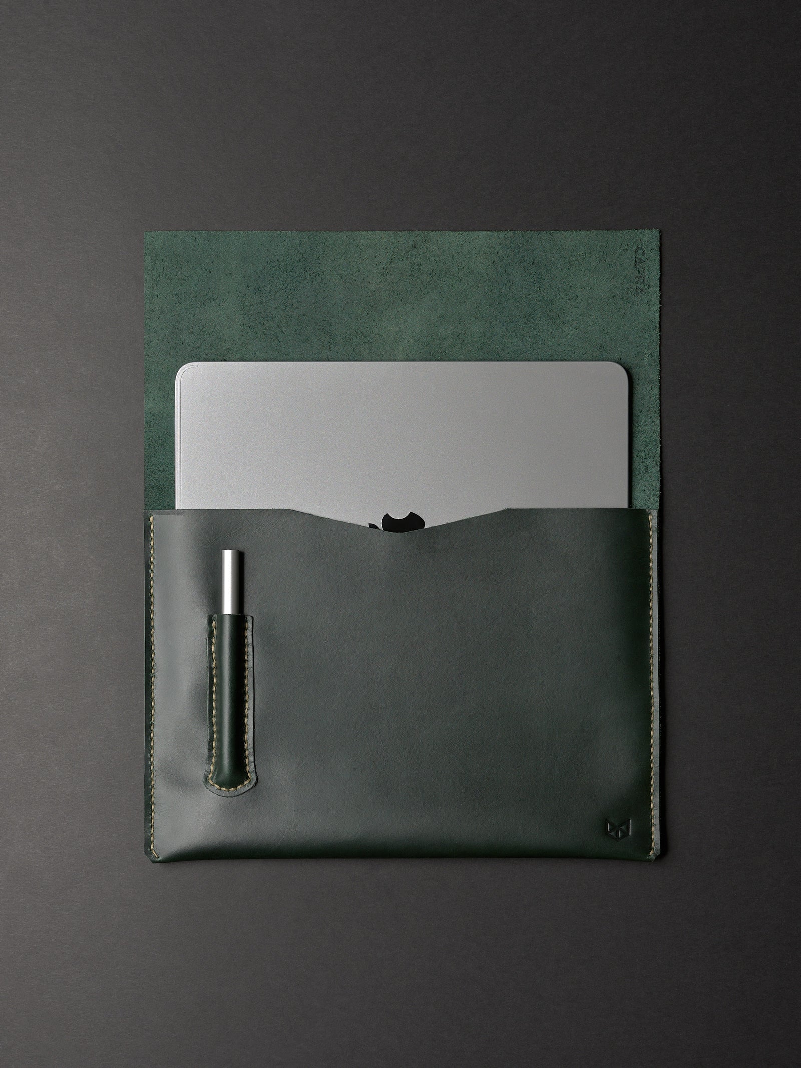 An open forest green leather sleeve partially holding a MacBook and a silver pen in a dedicated slot.