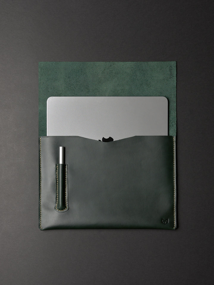 An open forest green leather sleeve partially holding a MacBook and a silver pen in a dedicated slot.