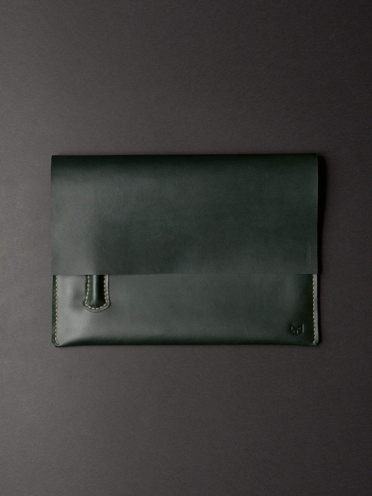 An overhead view of a closed, forest green leather laptop sleeve with a subtle logo in the corner on a dark background.