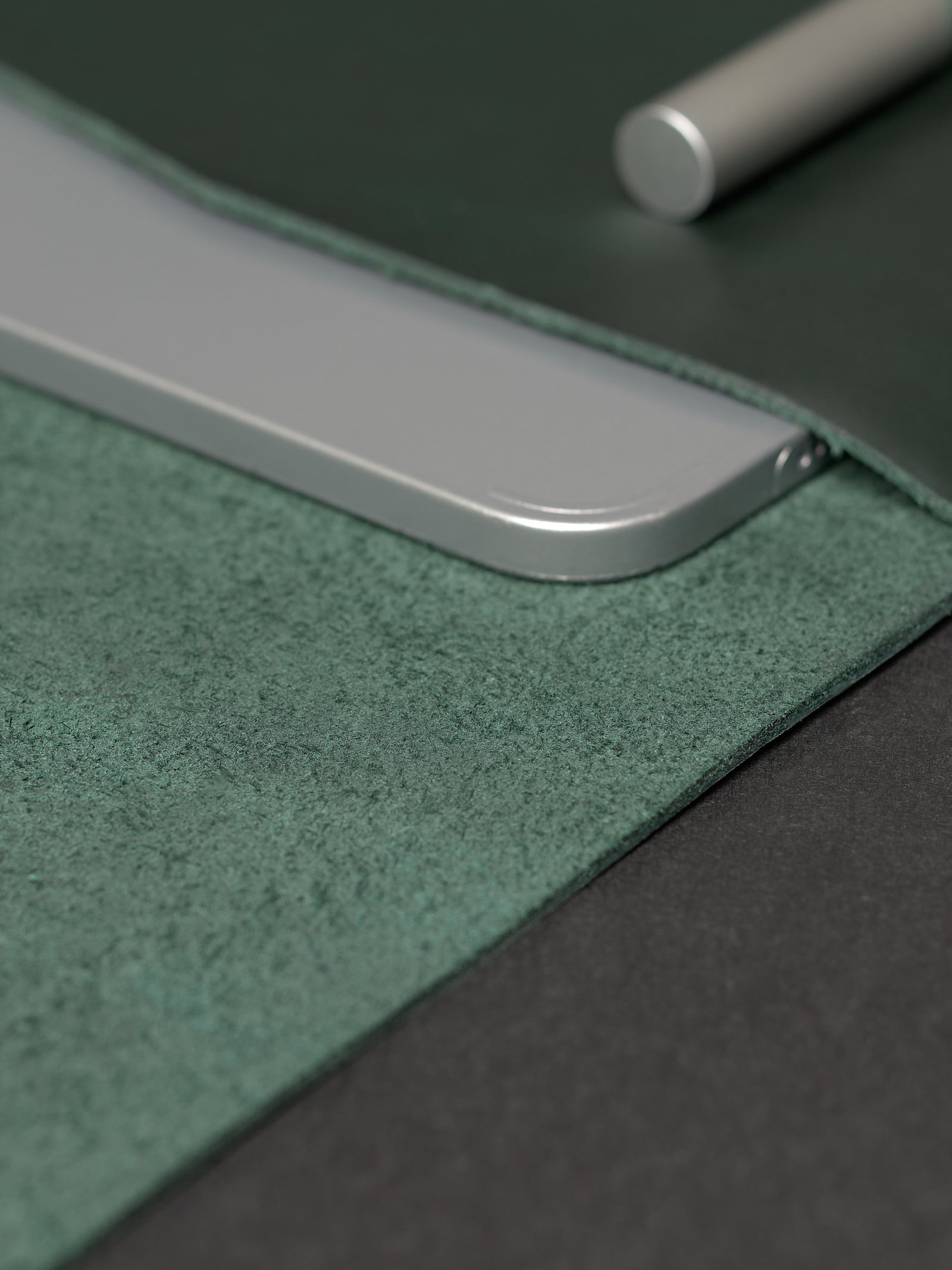 An overhead view of an open forest green leather laptop sleeve, pen, silver notebook, and aluminum ruler.