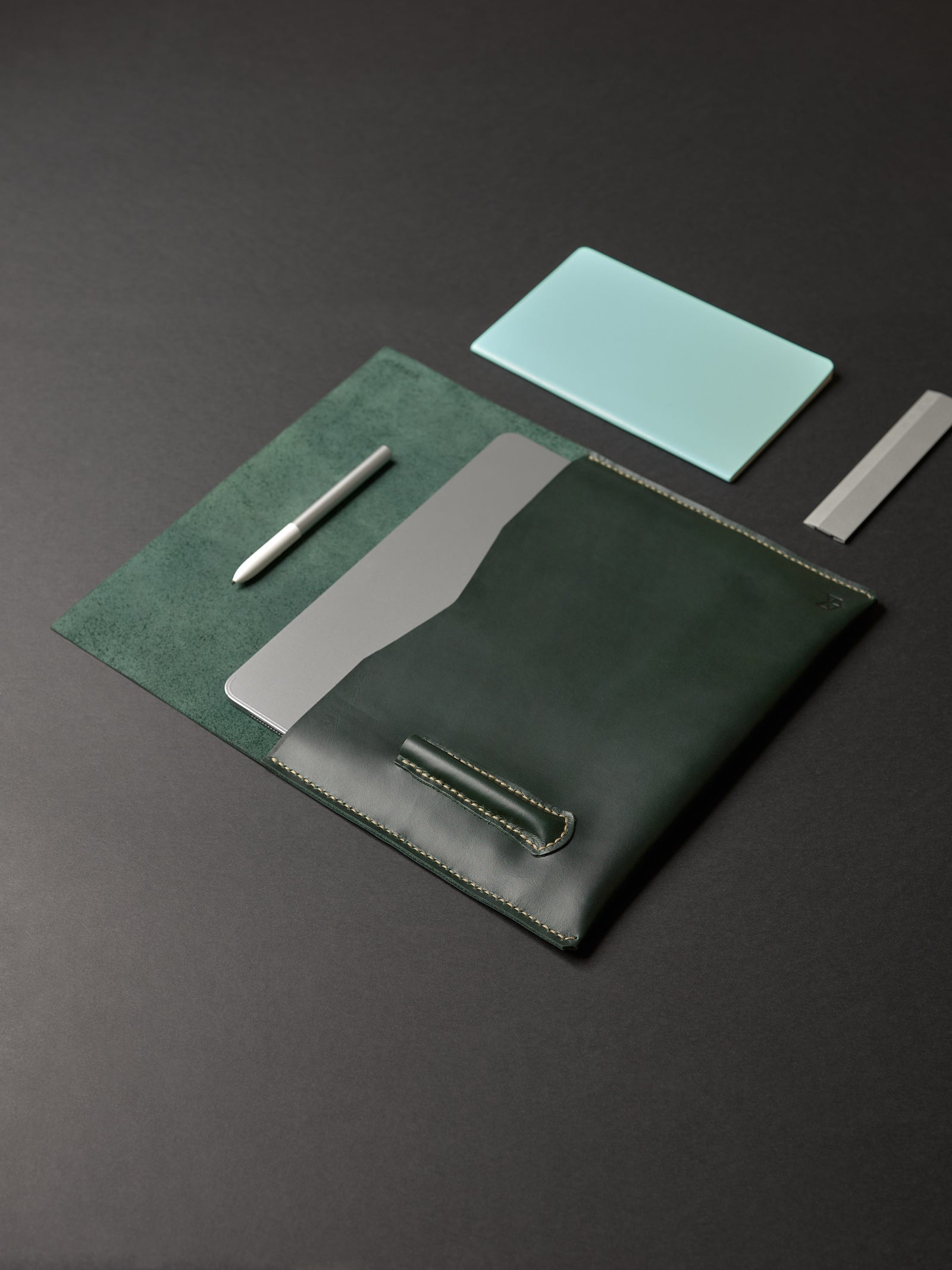 An overhead view of an open forest green leather laptop sleeve, pen, silver notebook, and aluminum ruler.