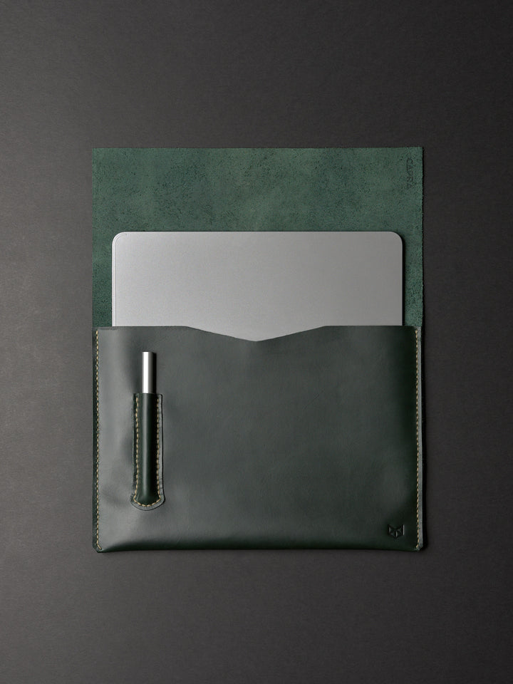 An overhead view of an open forest green leather laptop sleeve holding a silver laptop and a pen in a dedicated slot.