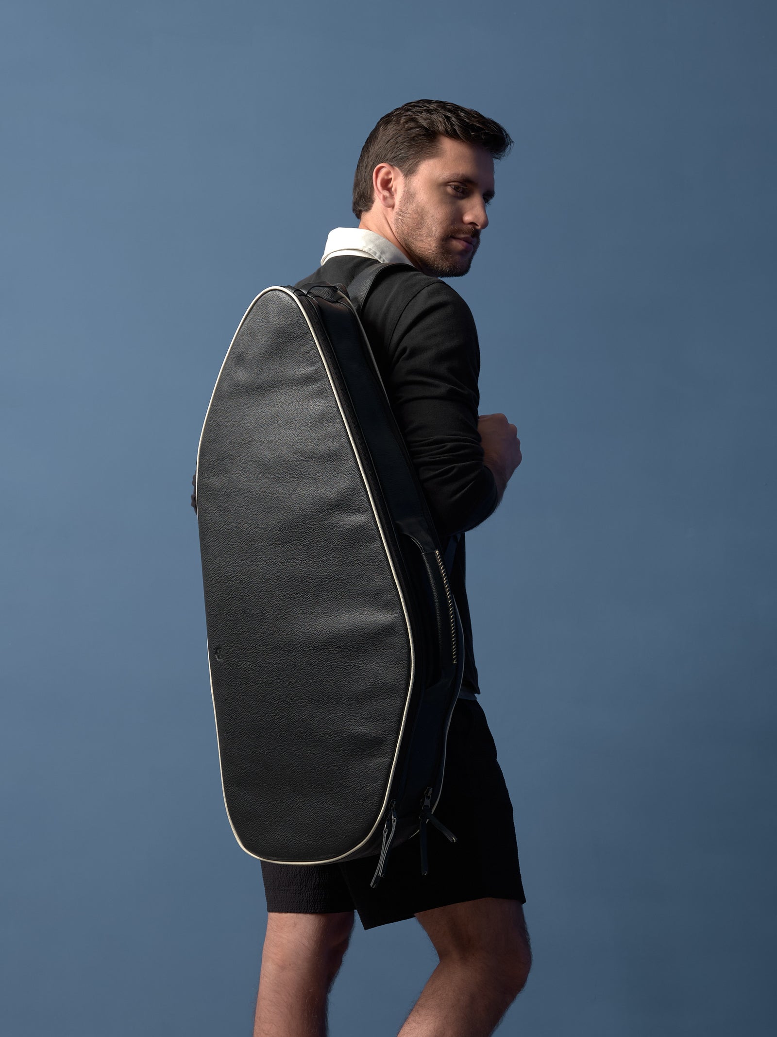 Man wearing a premium black leather tennis backpack with cream piping against a blue background.