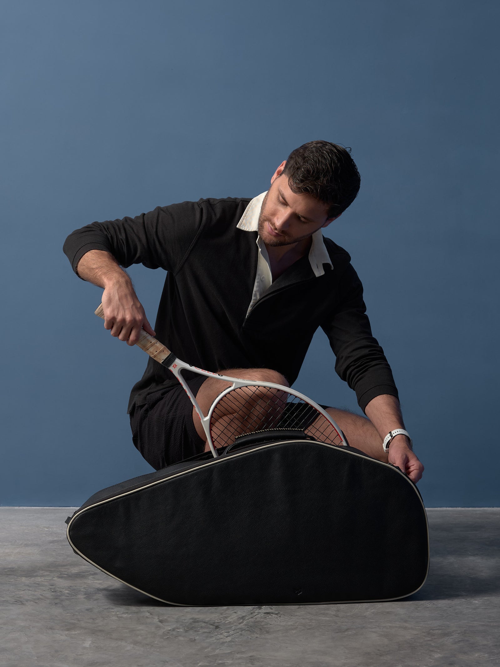 Man sliding a tennis racquet into a sleek, black full-grain leather tennis bag with cream trim.