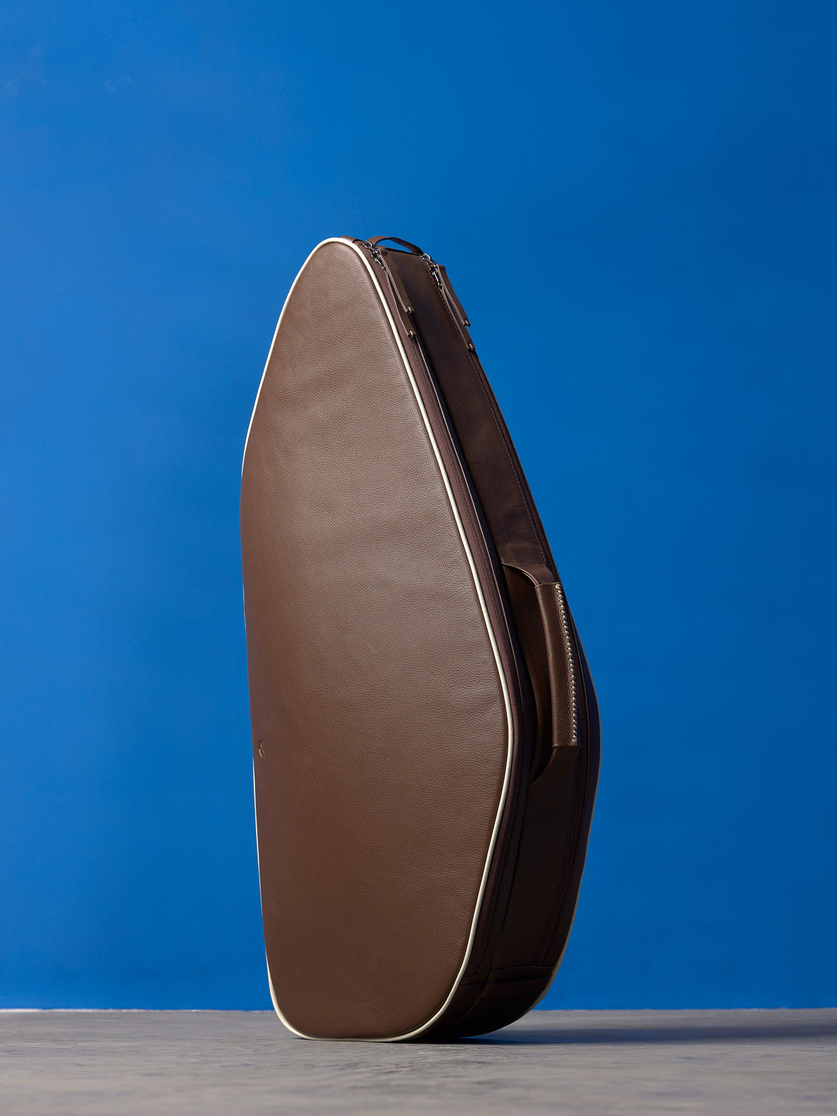 Vertical front view of a premium brown leather tennis bag with white piping against a blue background.