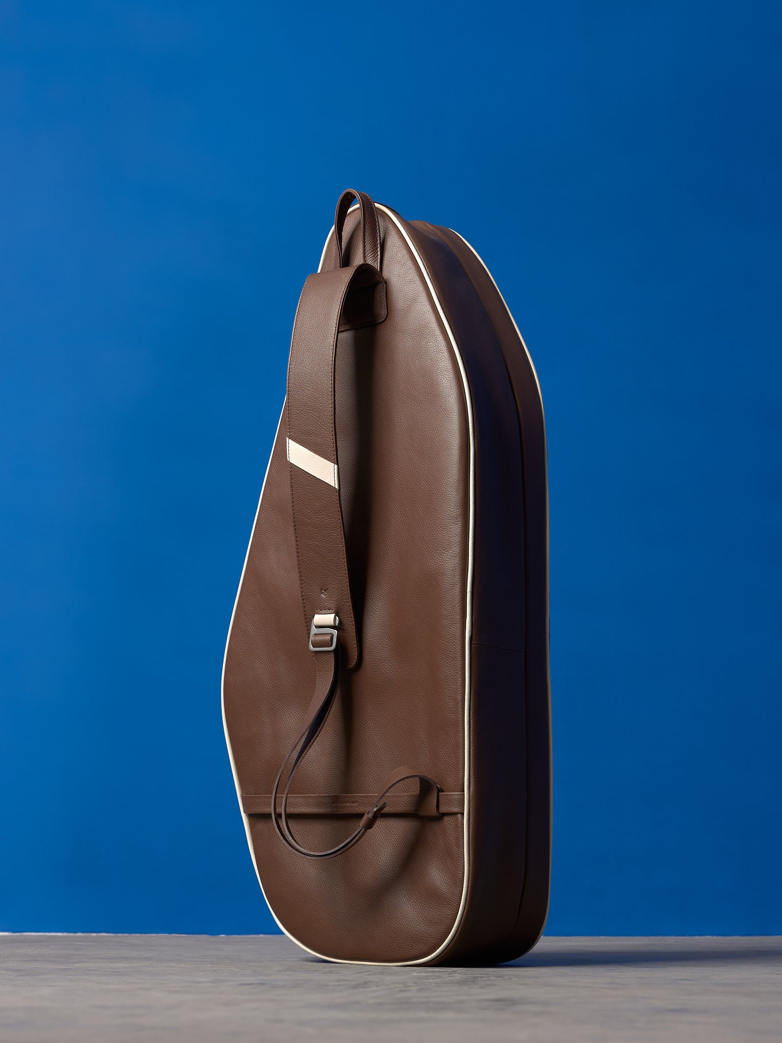 Rear view of a standing brown leather tennis bag, showcasing the single-strap backpack design and white piping.