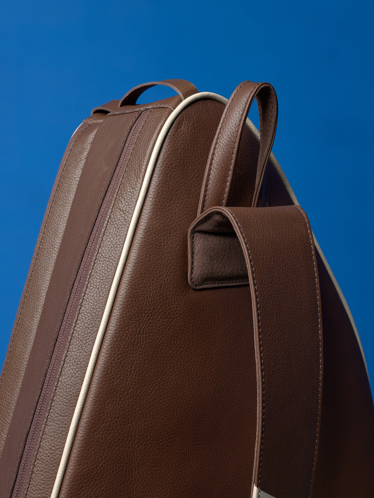Close-up of the top handle and reinforced stitching on a handcrafted leather tennis bag against a blue background.