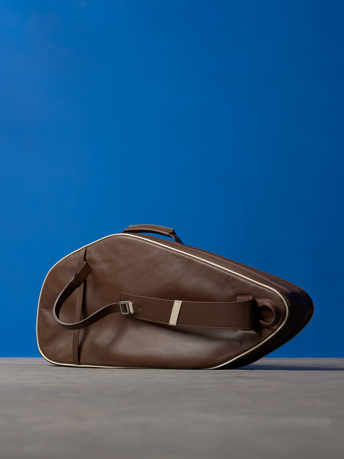 Side profile of a brown leather tennis racket bag laying flat, showing the adjustable shoulder strap and hardware.