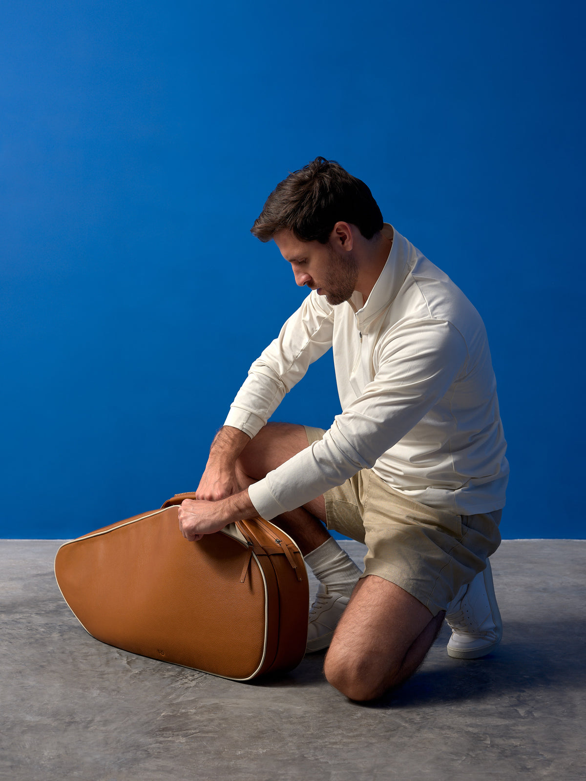 A man kneeling to open a luxury tan leather tennis bag, showcasing its size and soft-grain texture.