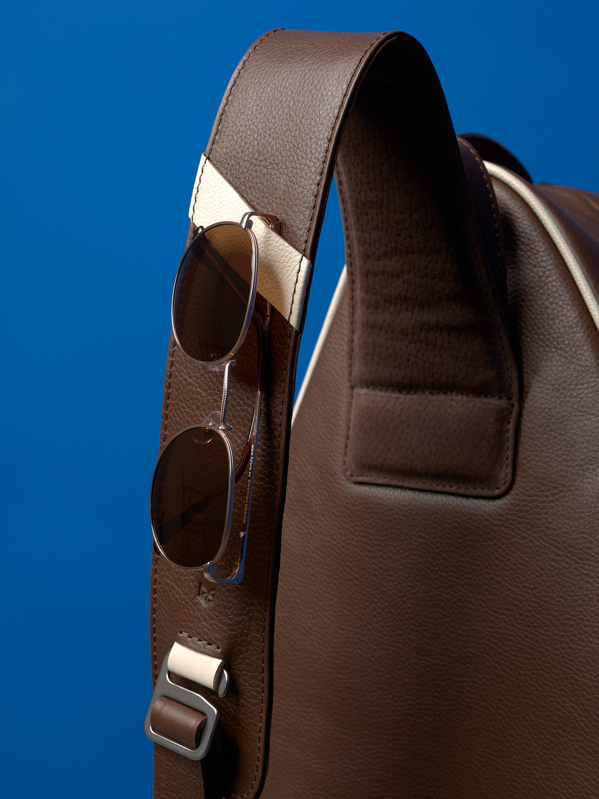Close-up of a leather bag strap featuring a dedicated sunglasses holder and contrasting cream leather accents.