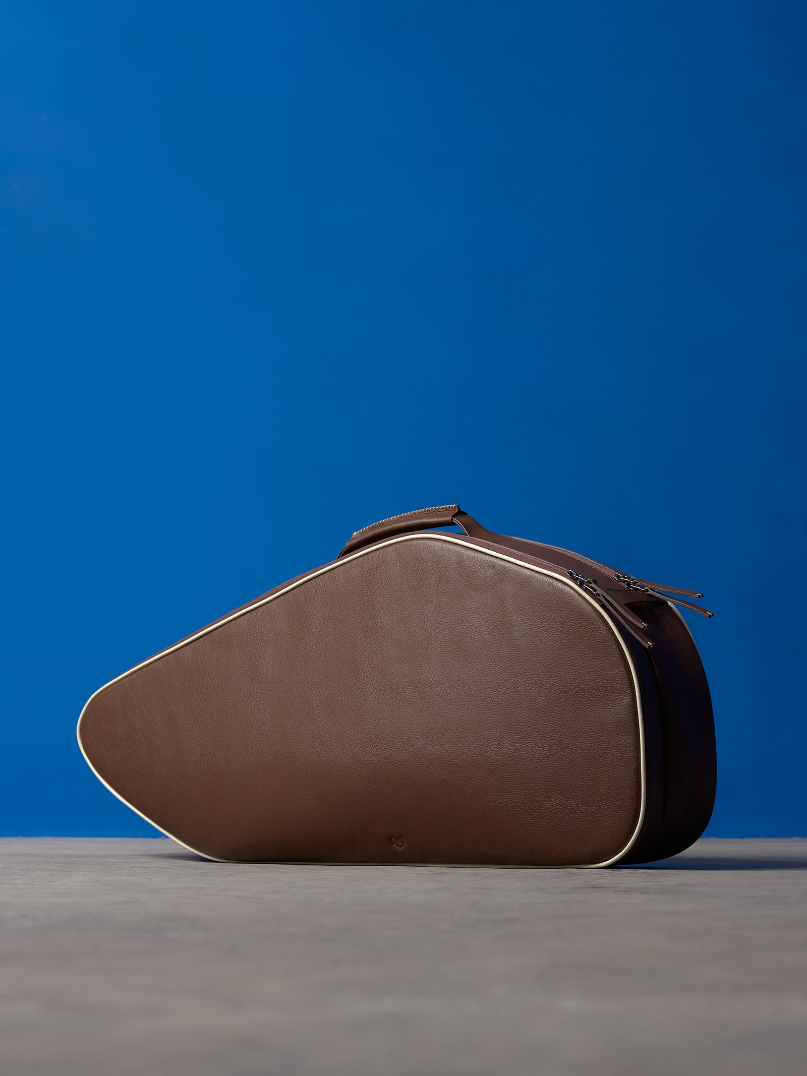 Horizontal view of a closed brown leather tennis bag on a concrete surface against a minimalist blue backdrop.