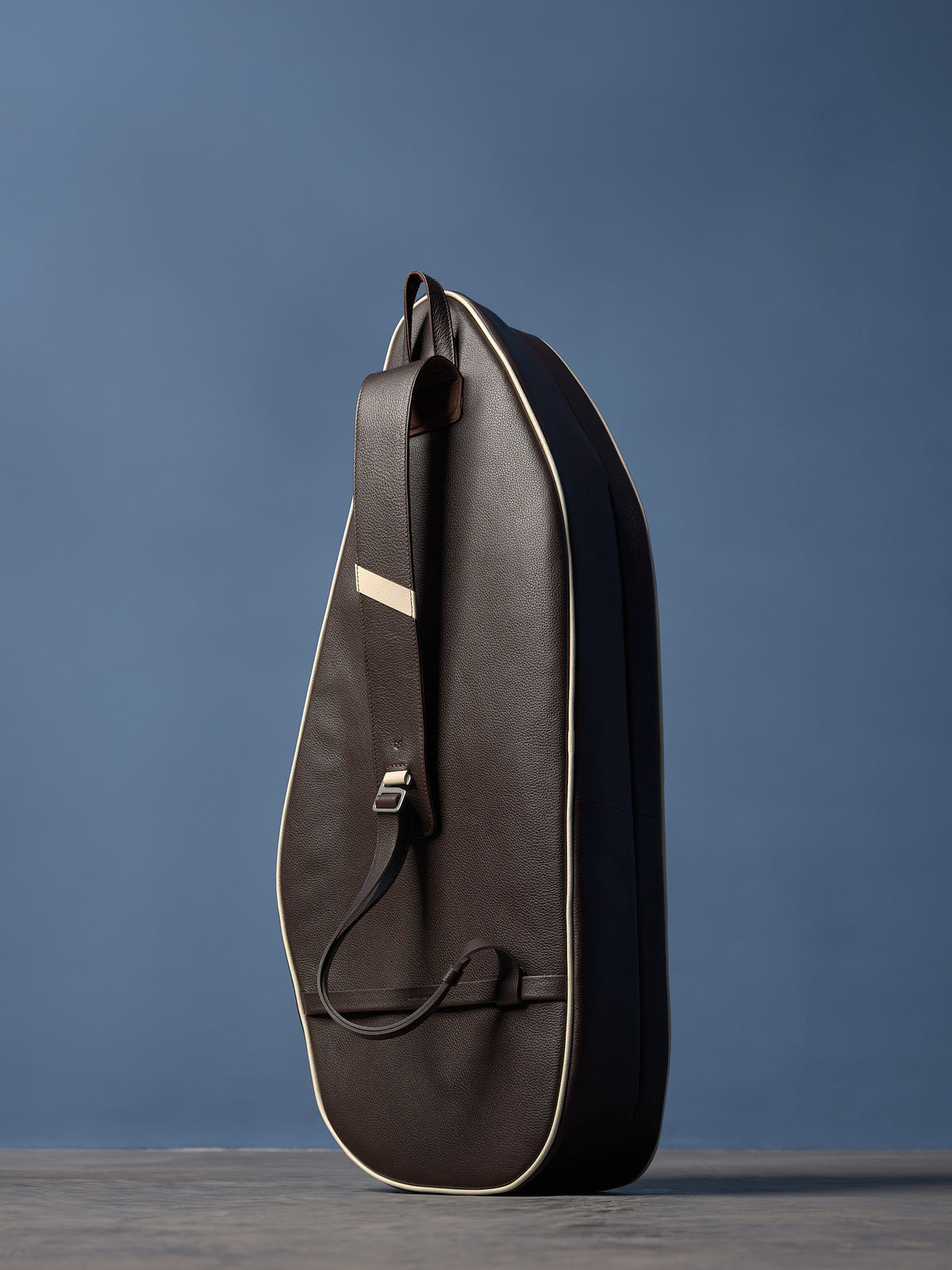 Dark brown pebbled leather racket bag with cream piping, single shoulder strap, and polished buckle, standing upright.
