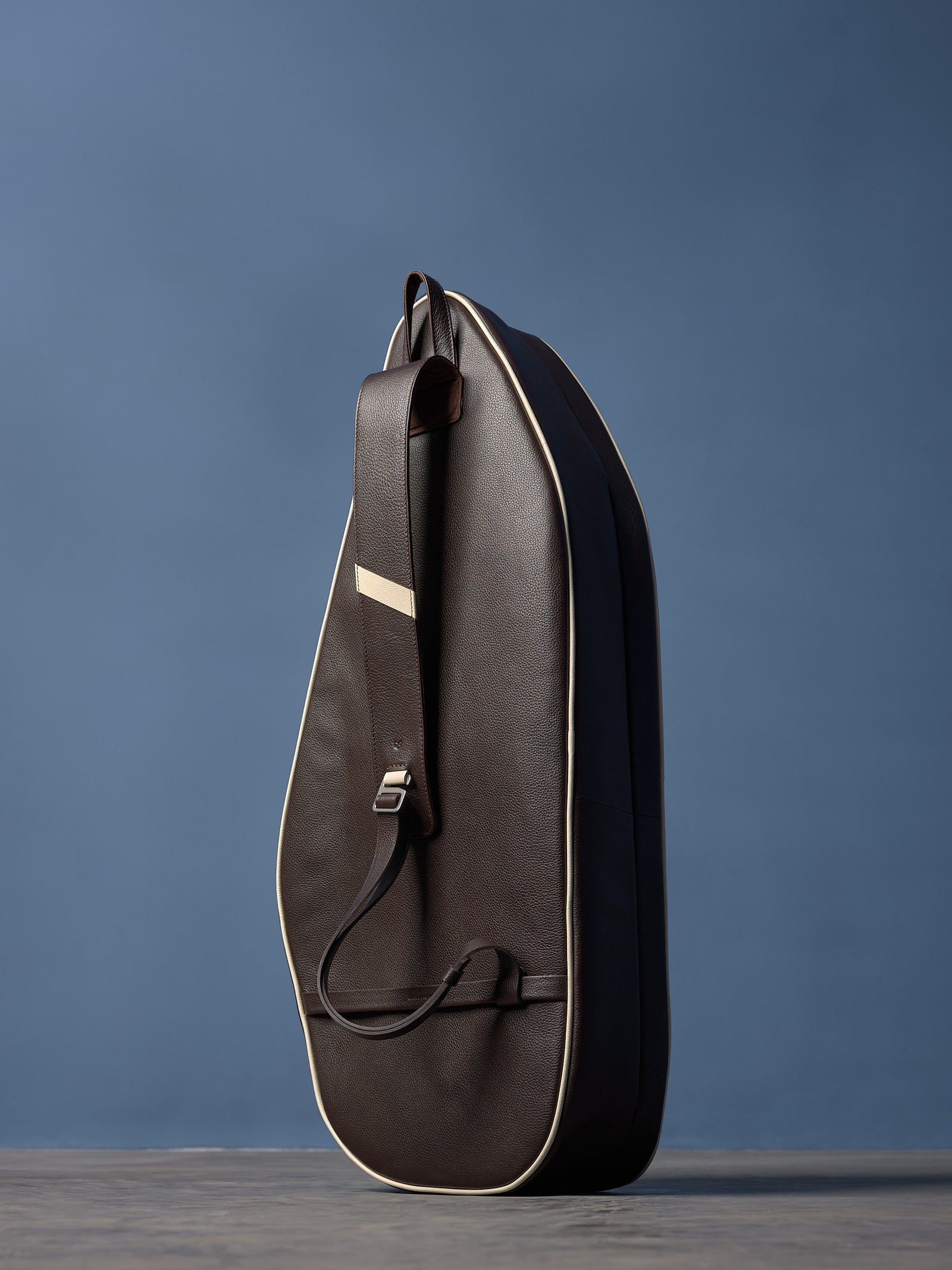 Dark brown pebbled leather racket bag with cream piping, single shoulder strap, and polished buckle, standing upright.