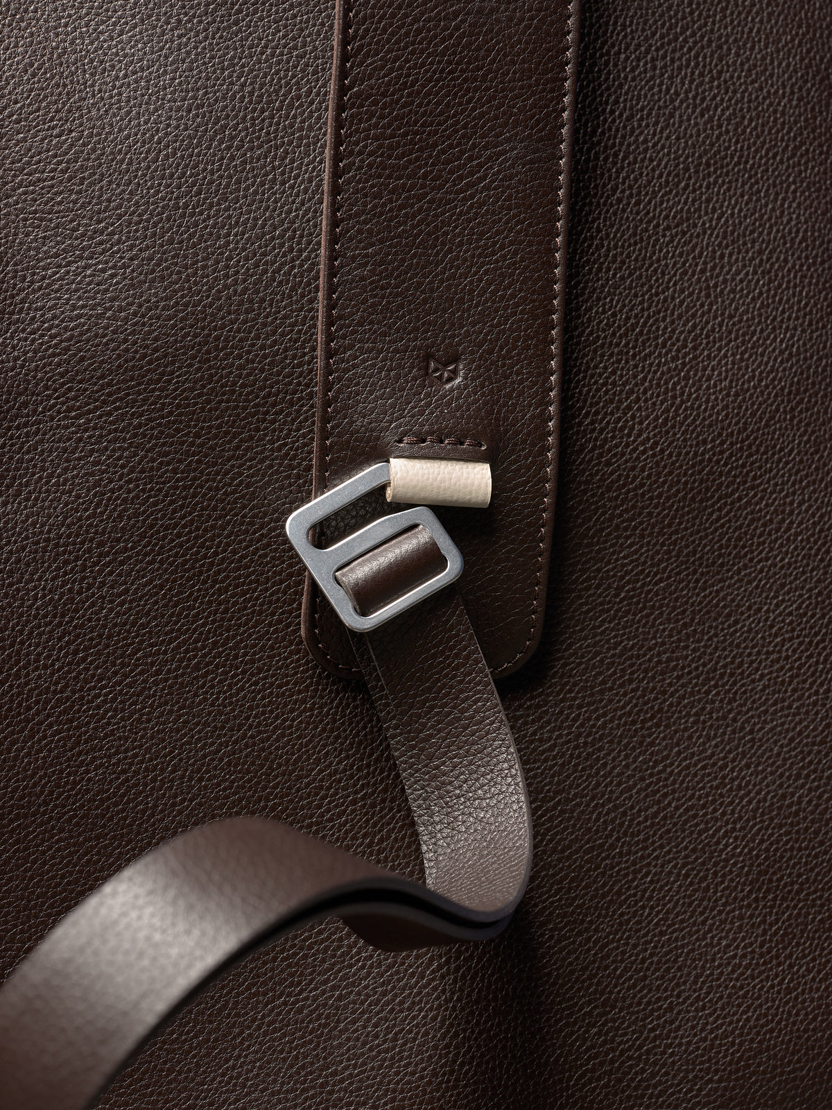 Close-up of a dark brown pebbled leather bag, featuring a metallic buckle, a leather strap, and a subtle stamped logo.