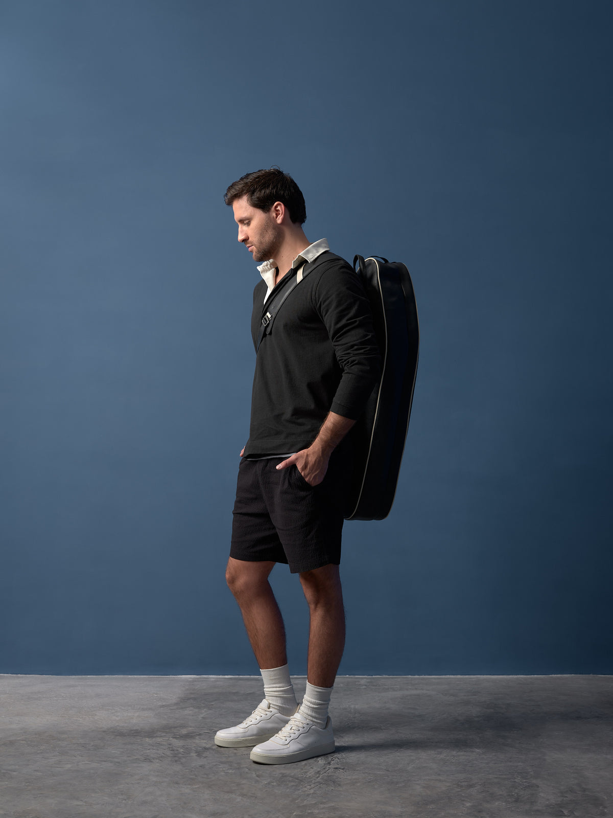 Male model in black shirt and shorts, with white socks and sneakers, casually carries a black tennis bag on his shoulder.