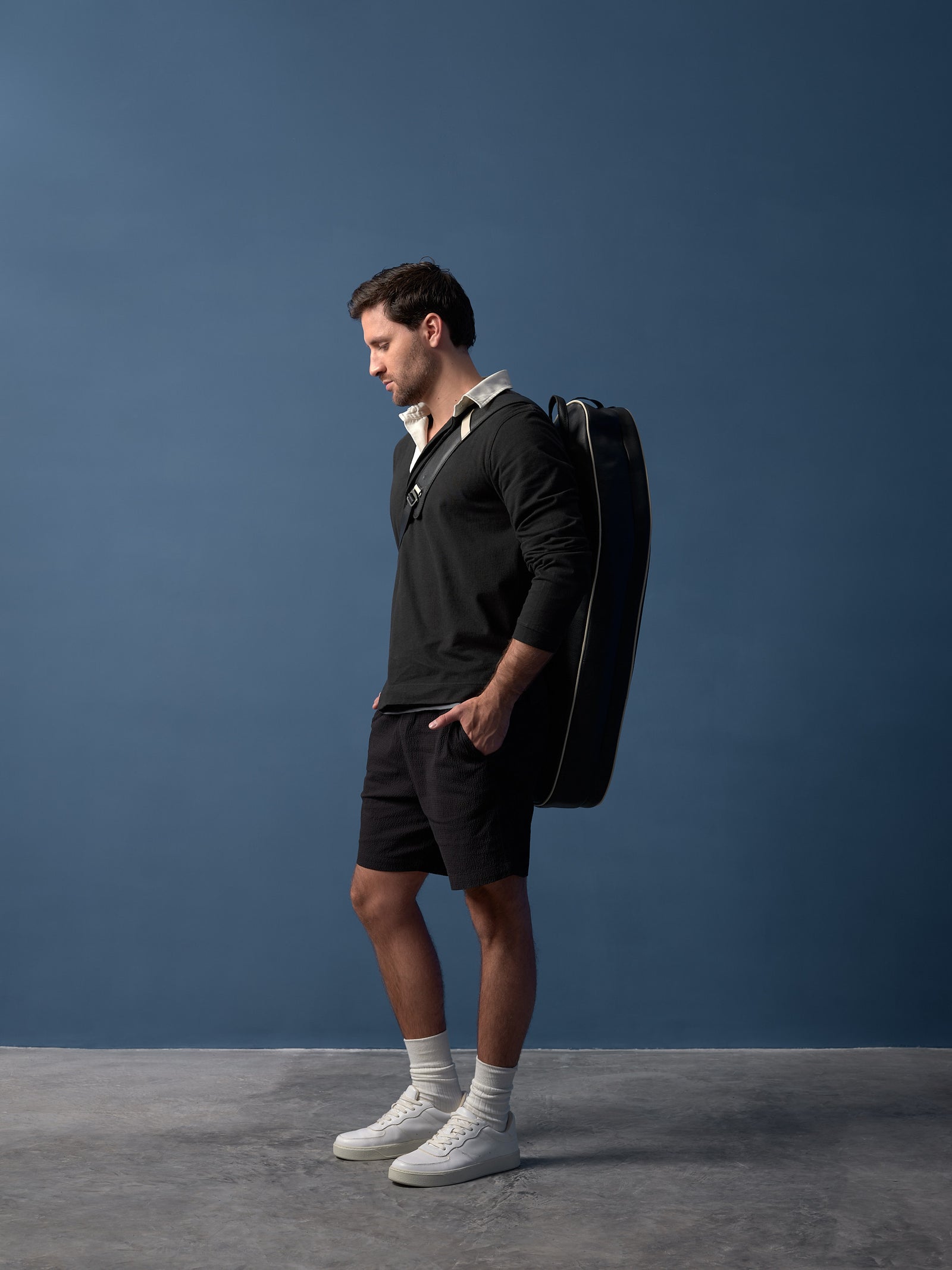 Male model in black shirt and shorts, with white socks and sneakers, casually carries a black tennis bag on his shoulder.