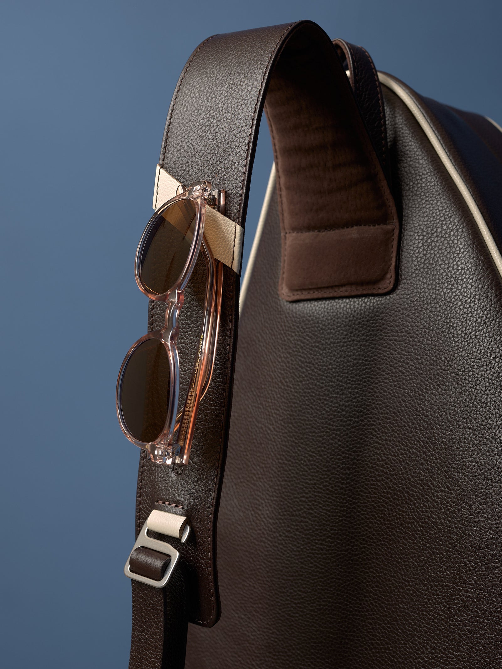 Detail shot of brown leather racket bag strap with polished buckle and a pair of modern, round clear-framed sunglasses.