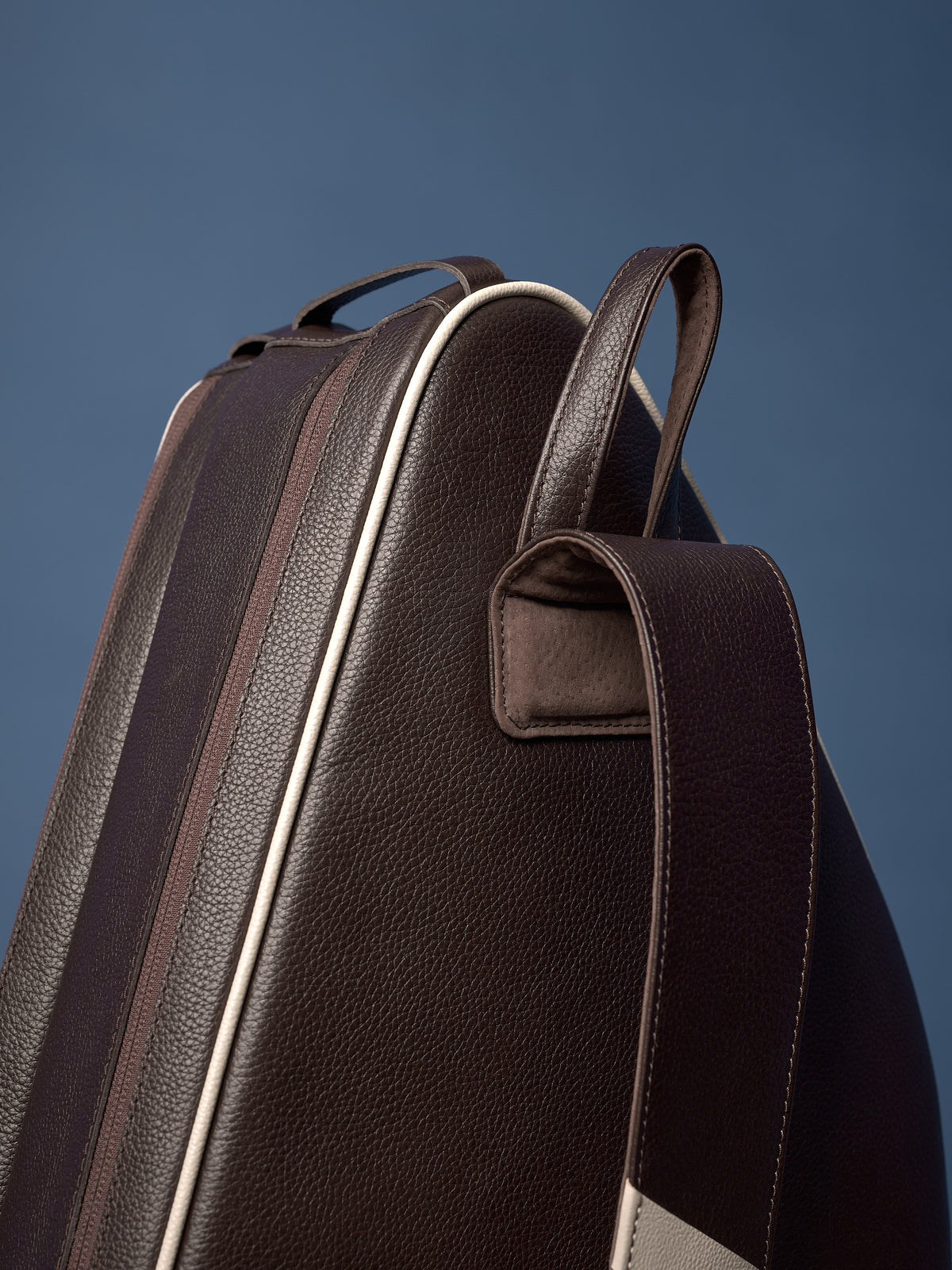 Angled view of dark brown pebbled leather bag, showcasing cream piping, sturdy loop handle, and padded shoulder strap attachment.