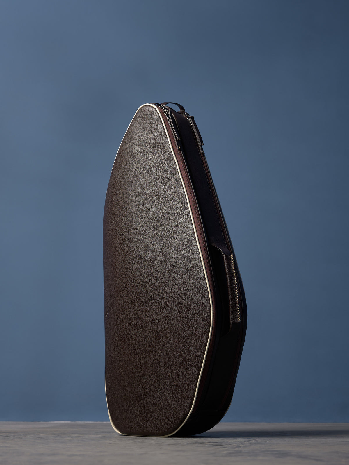 Side profile of dark brown textured leather racket bag with distinct cream piping and dual main compartment zippers.