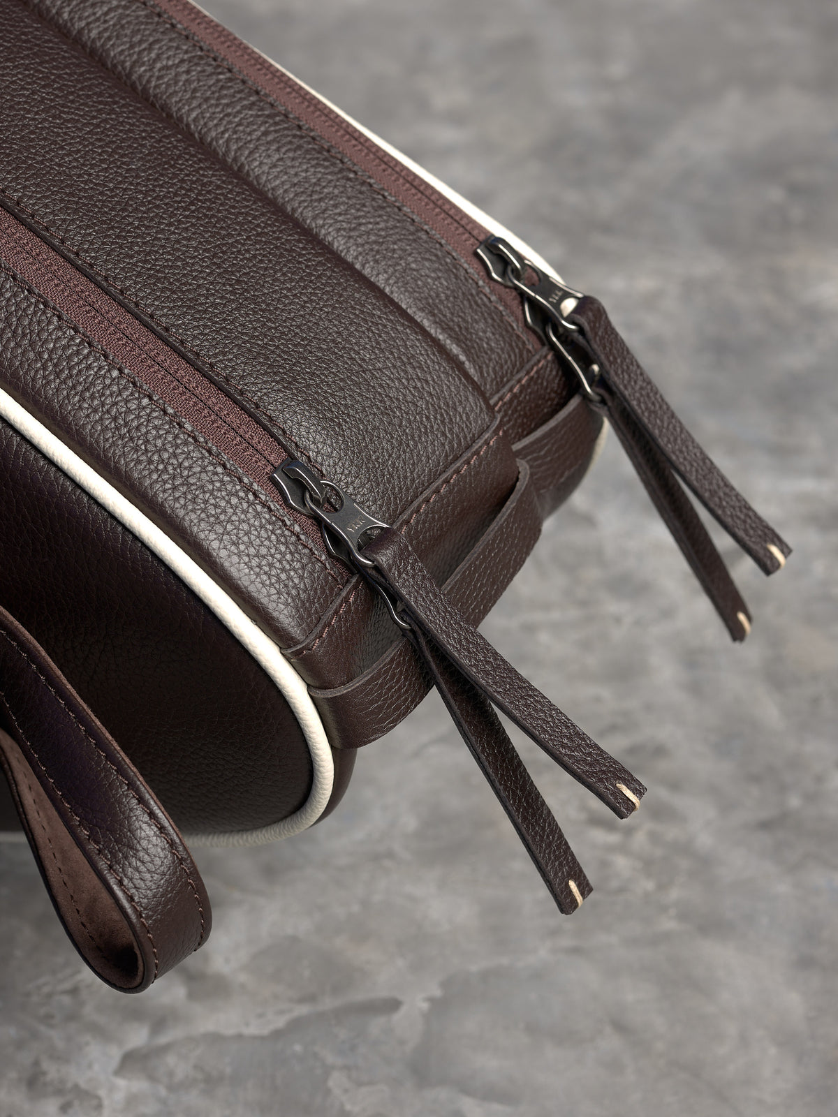 Macro detail of premium leather-pull zippers on a dark brown bag, highlighting the pebble grain and precise cream stitching.