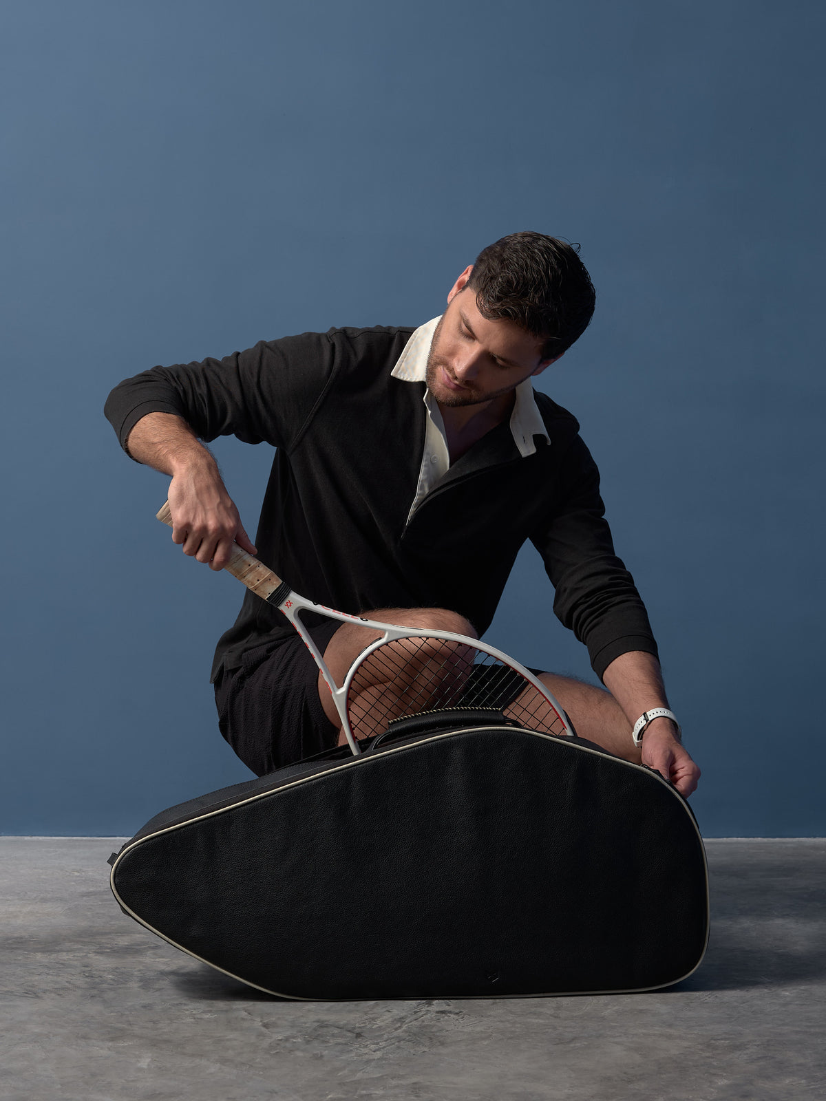 A man in a black polo shirt, black shorts, and white sneakers kneeling to carefully place a white tennis racket into a brown leather Capra Leather bag.