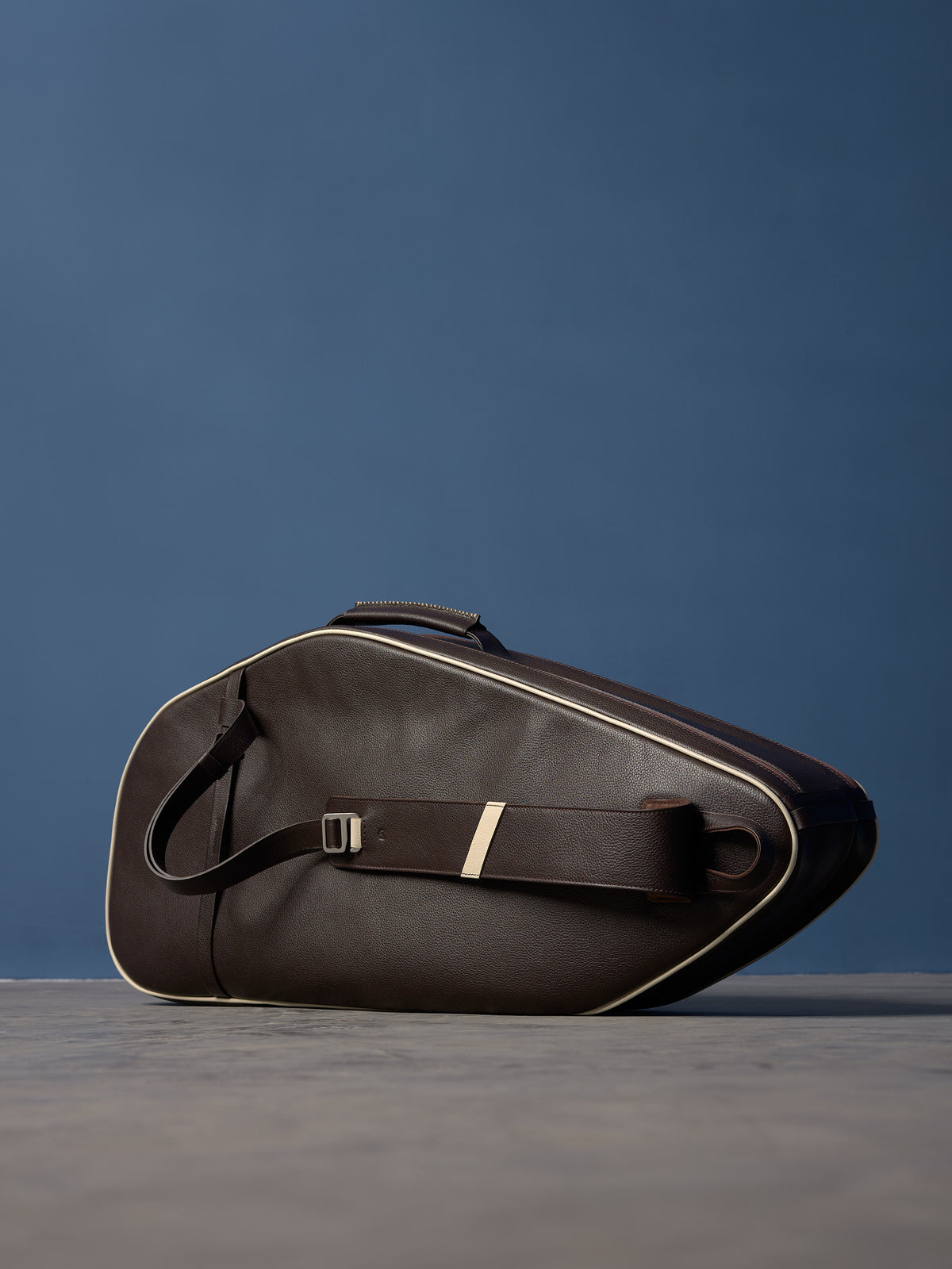 Side view of a dark brown leather racket bag, with the leather and cream-striped shoulder strap resting across the main body.