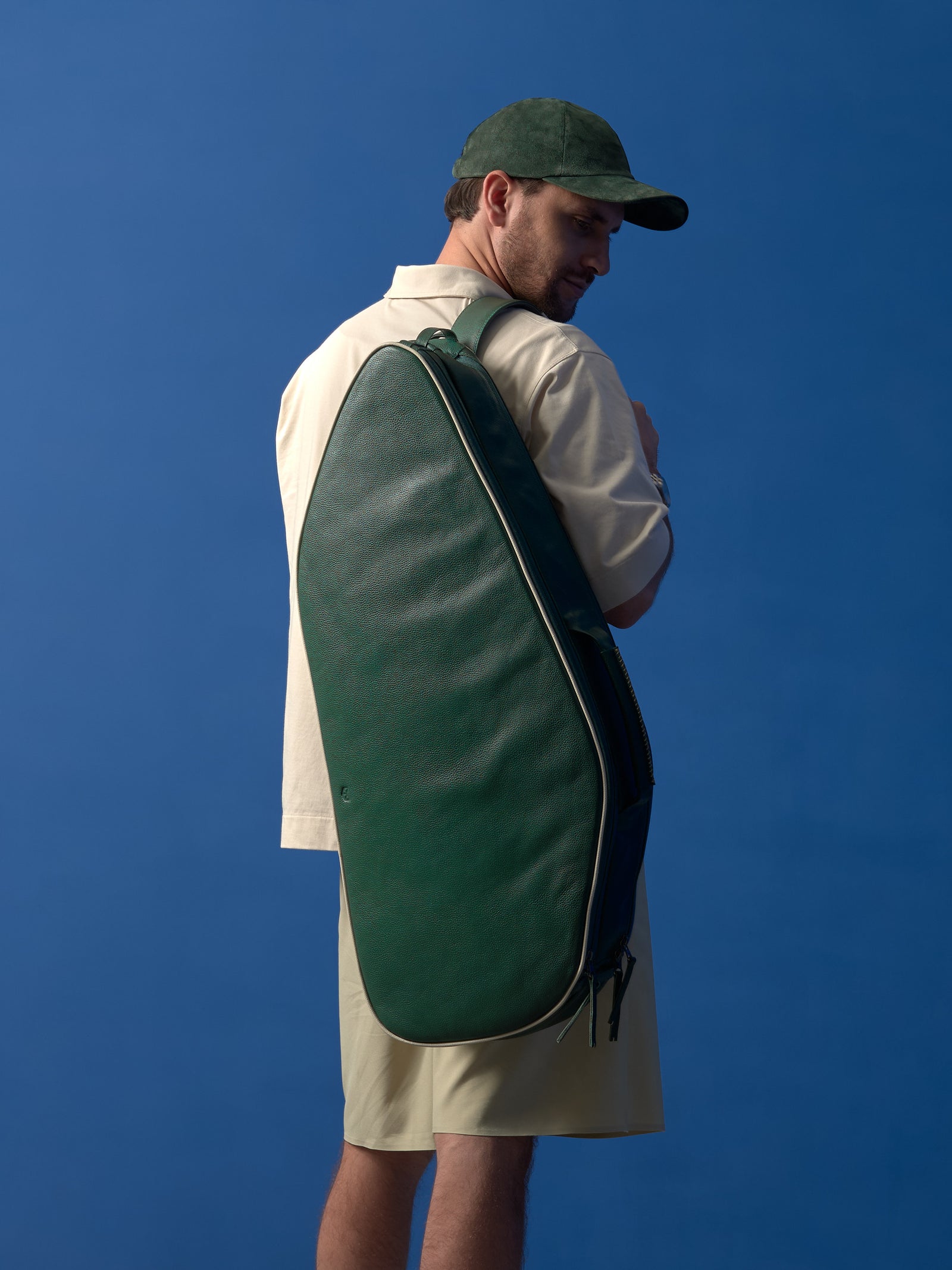 Rear view of a man carrying the dark green leather tennis bag over one shoulder, showcasing its ergonomic teardrop shape and the smooth full-grain leather finish.