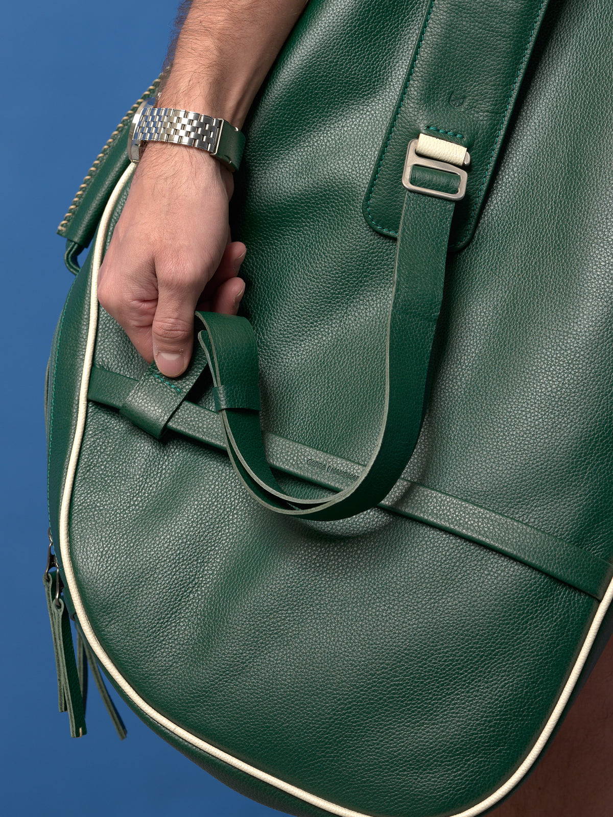 A close-up of the adjustable leather strap being tucked into a matching green leather keeper on the side of the bag, emphasizing the minimalist hardware and construction.