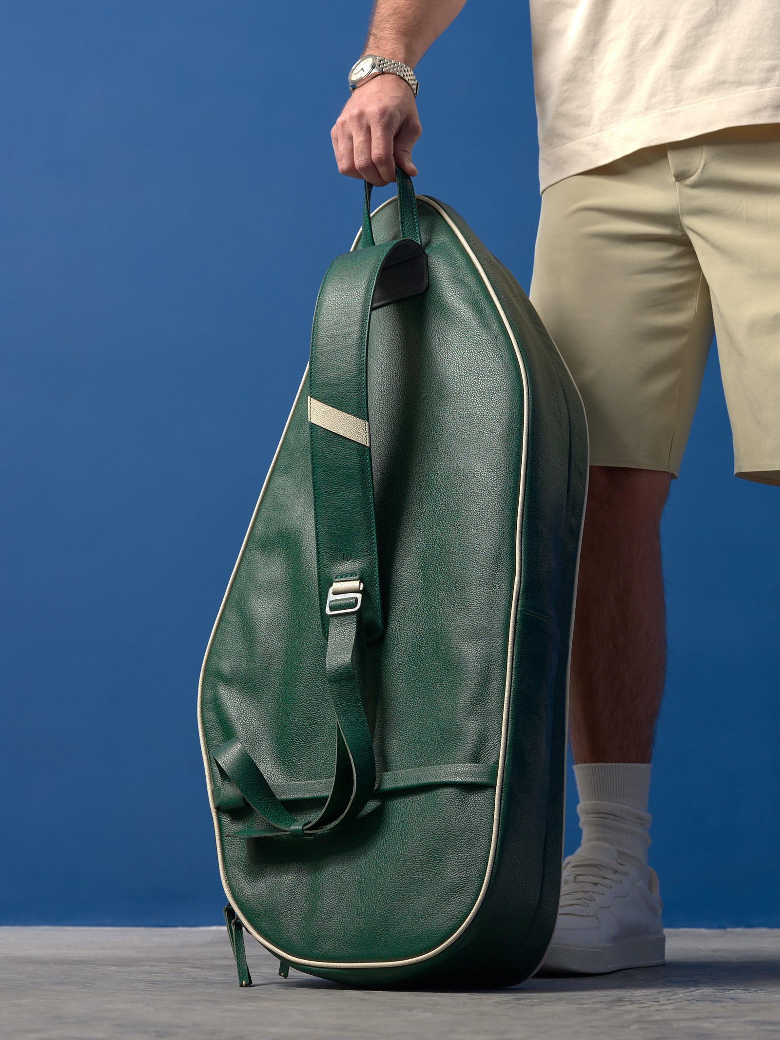 A full-body lifestyle shot of a man standing next to a classic wooden director’s chair, holding the dark green leather tennis bag against a cobalt blue studio wall.